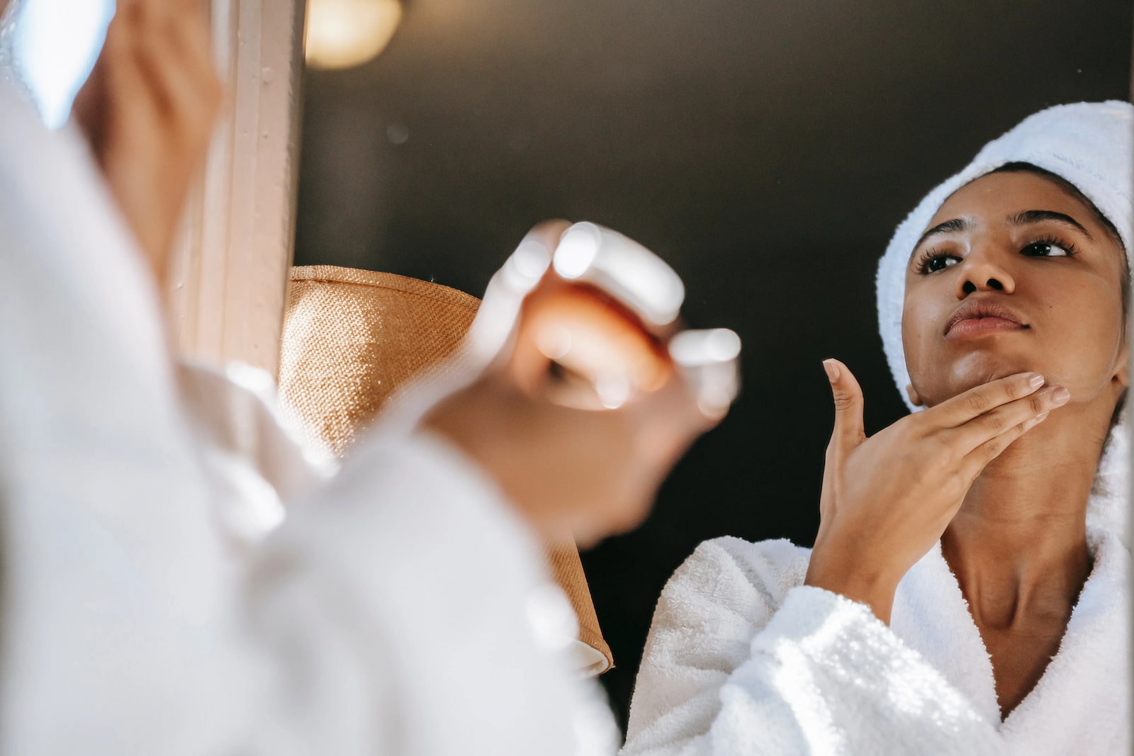 Woman in a white bathrobe and towel applying skincare product to her chin while looking in the mirror.