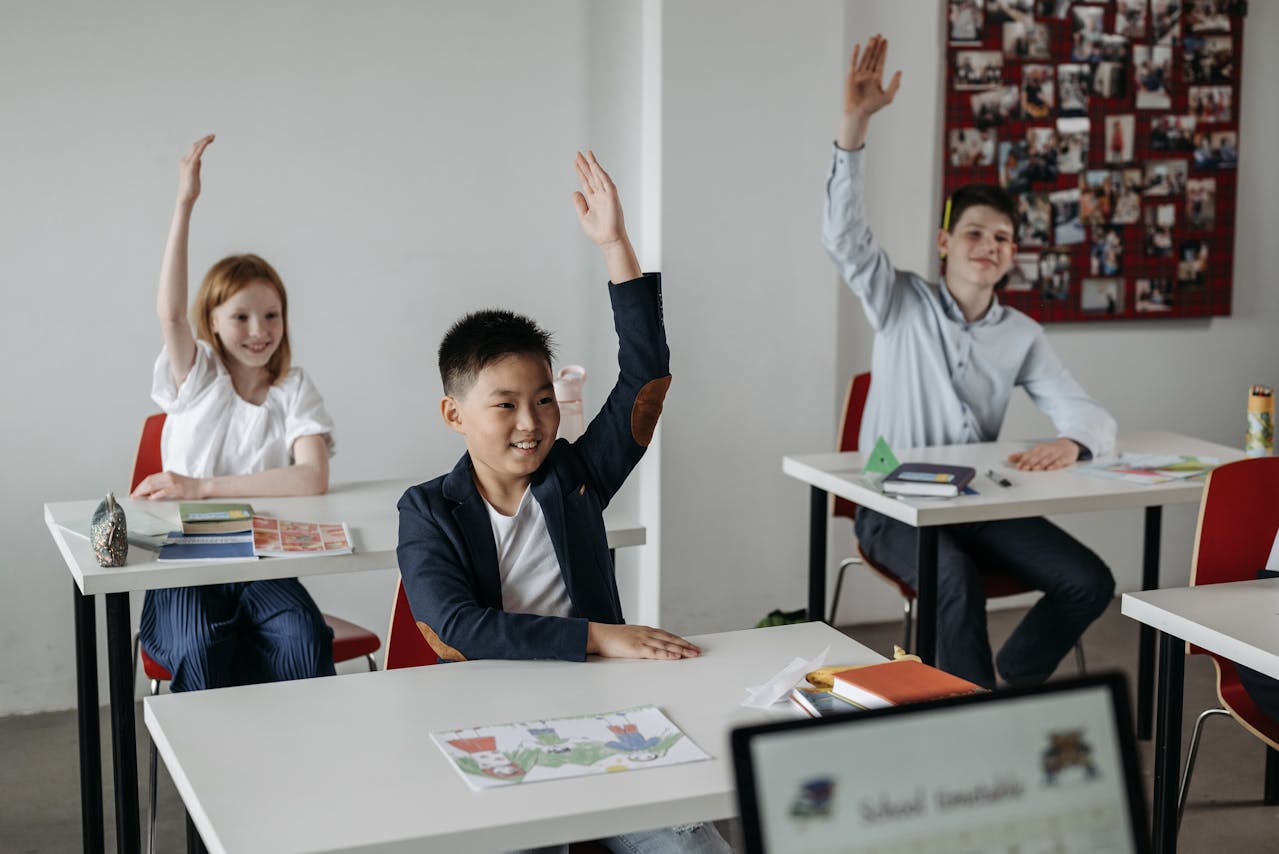 a bunch of happy students in a classroom, sitting at their desks and raising their hands 