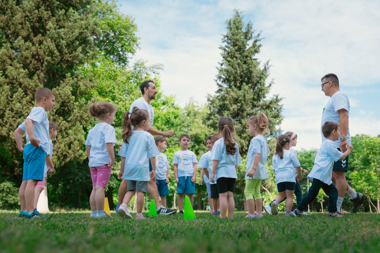 two teachers managing a group of young kids at a summer camp in a park