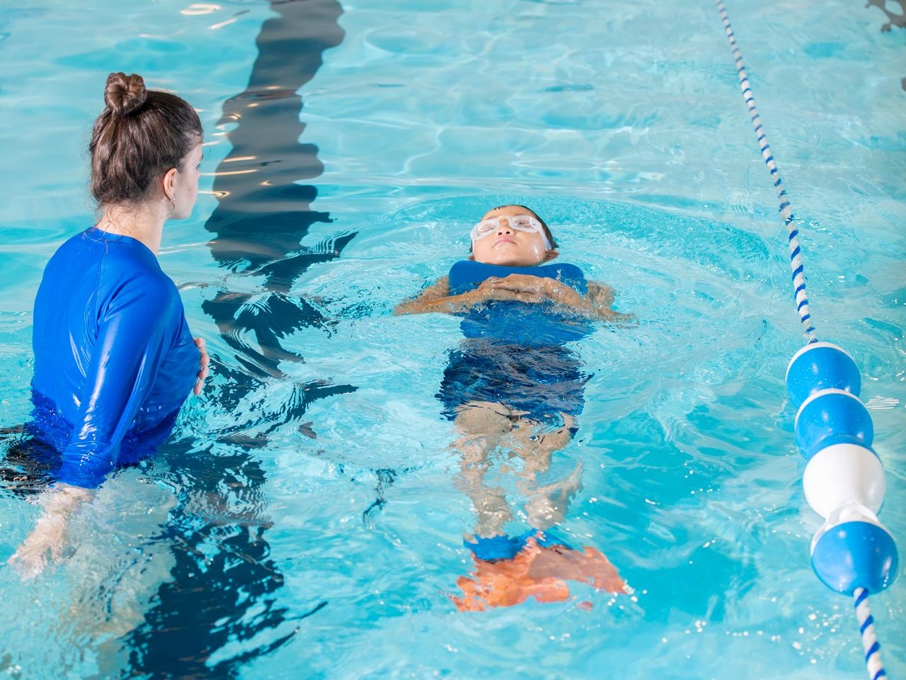 a swimming instructor coaching a boy floating on his back in the pool with a kickboard on this chest