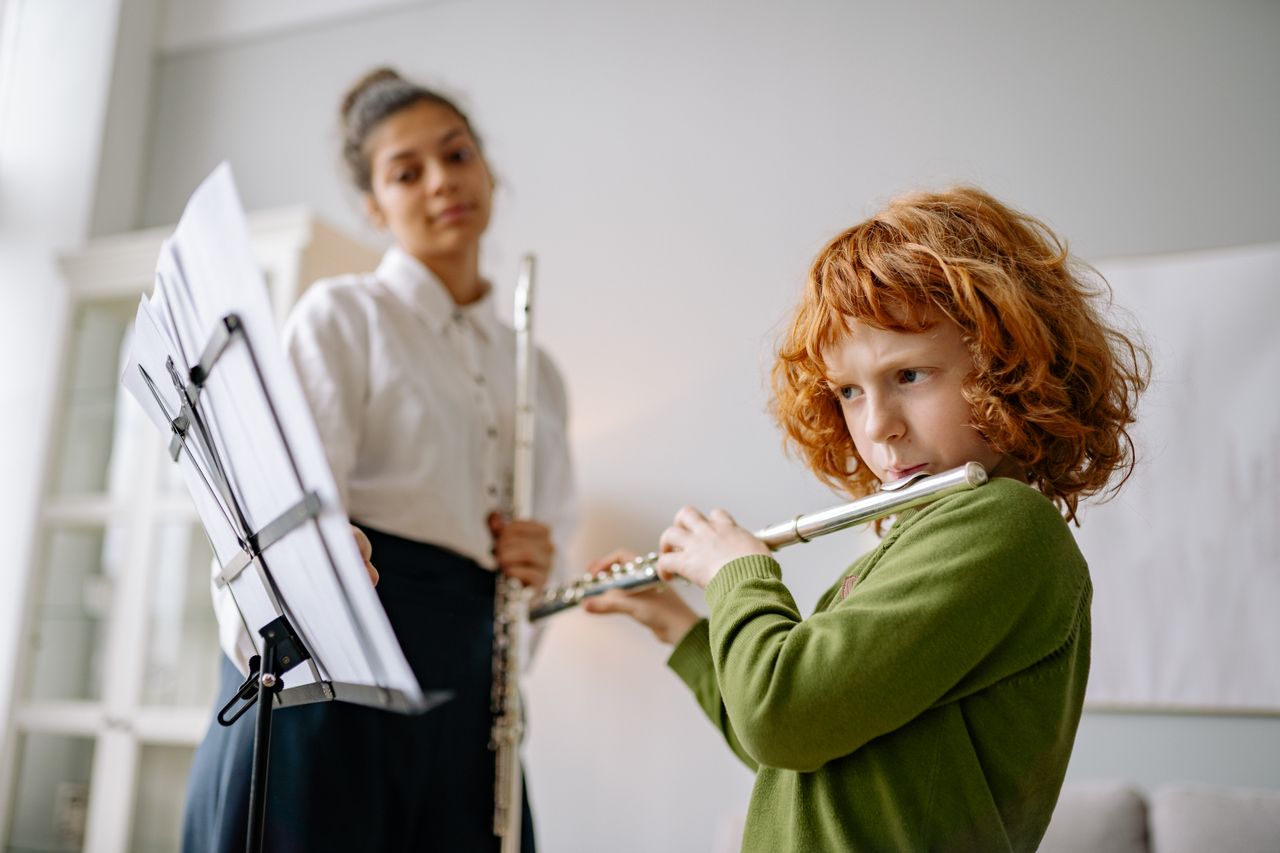 a young boy playing the flute while reading sheet music while his music teacher supervises him