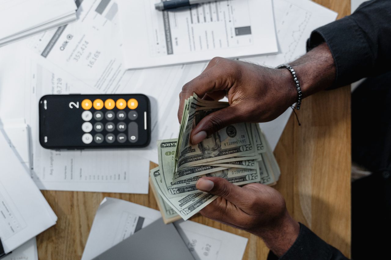 a man counting money, with papers strewn haphazardly on the table below him, and his mobile phone calculator showing a computation