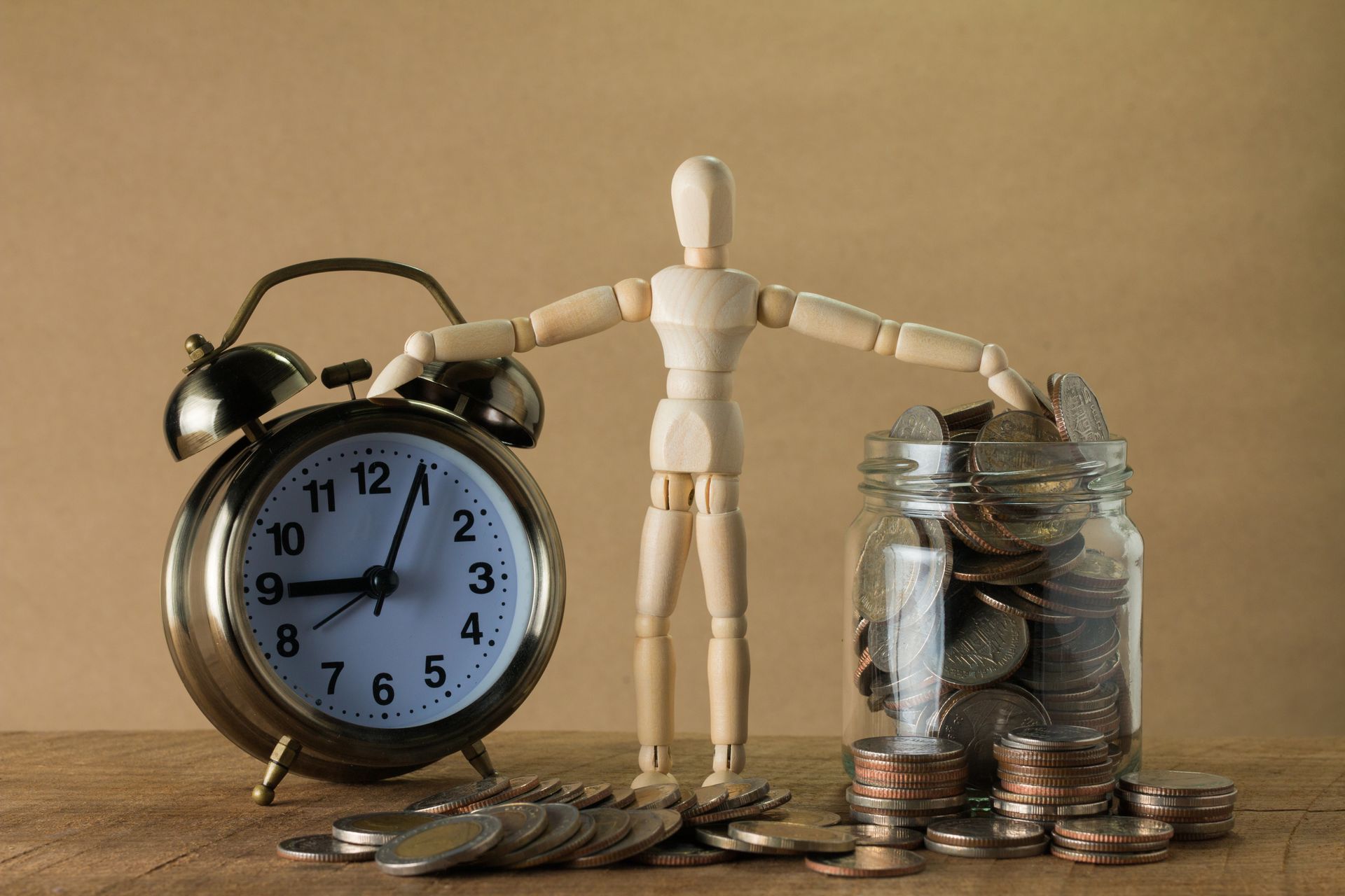 a small wooden mannequin touching an alarm clock with one hand and a jar of coins with the other, representing the concept of saving time and money