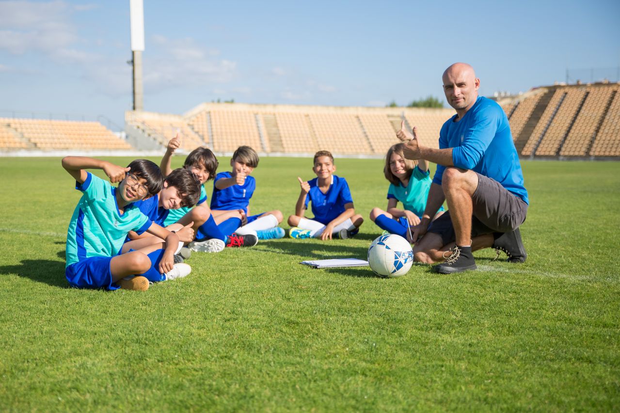 a coach and a group of young boys in football jerseys sitting on a football field, giving a thumbs-up and smiling at the camera