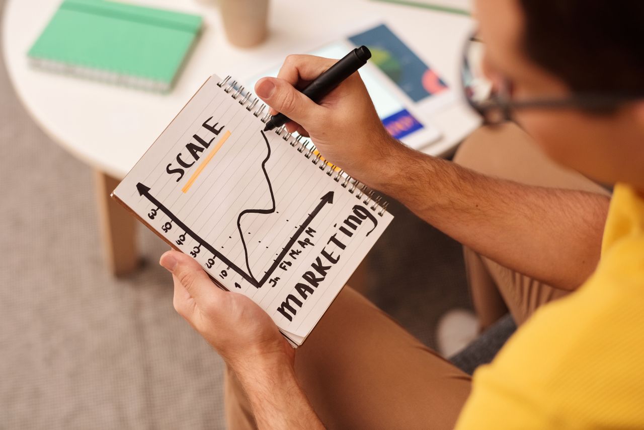 a man drawing a graph with a marker in his notebook, with the words 'Scale' and 'Marketing' written at the top and bottom of the page respectively