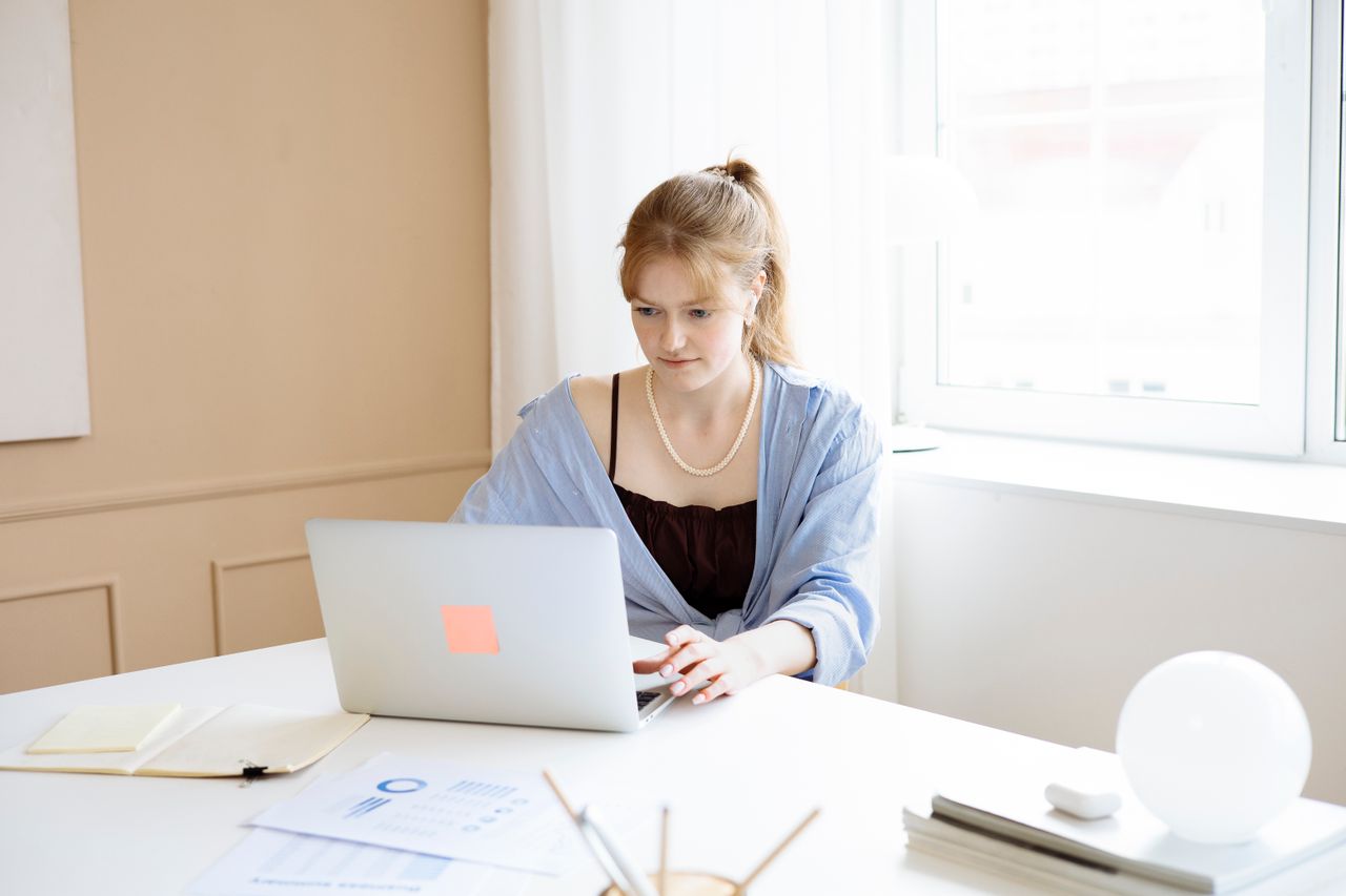 a young lady working on a laptop in a well-lit room, a notebook, some papers with graphs printed on them, and some stationery spread out across the table