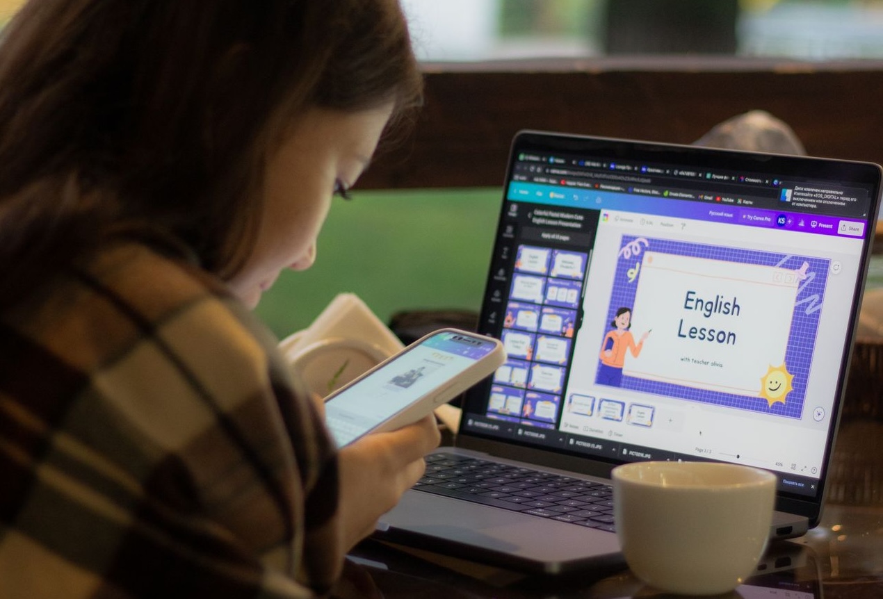 A woman using her laptop for an online English lesson while checking her phone.