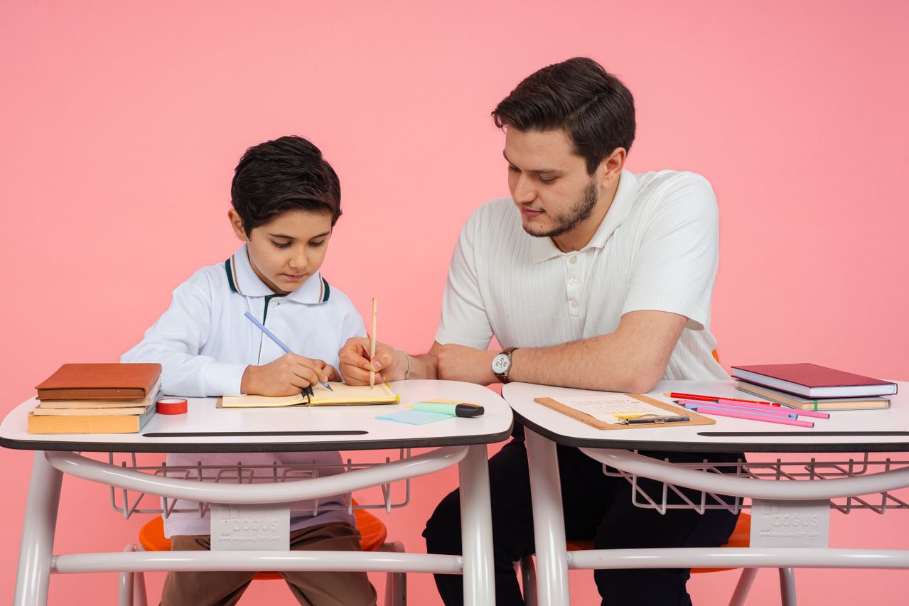 A teacher helps a student with schoolwork at a desk against a pink background.