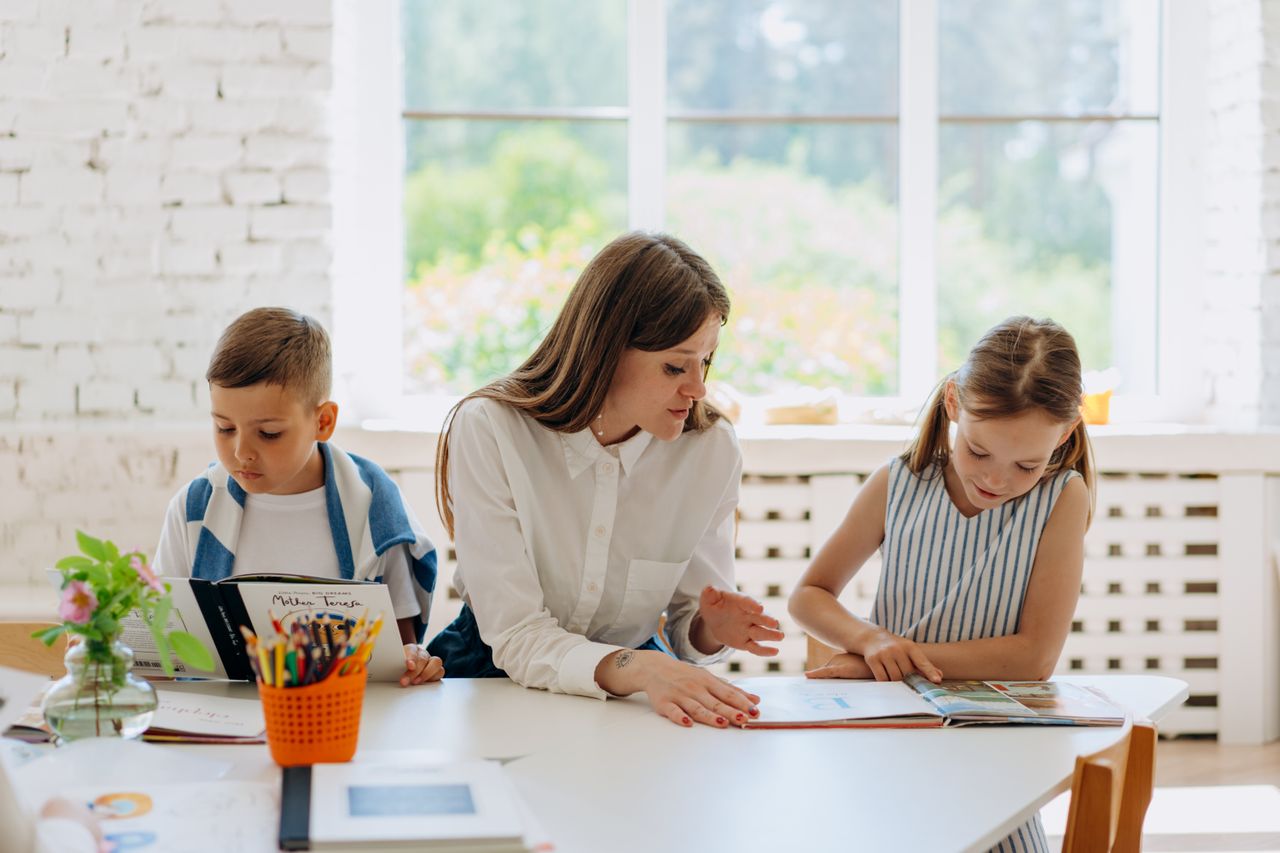 A female tutor helps two children learn by reading together in a bright classroom.
