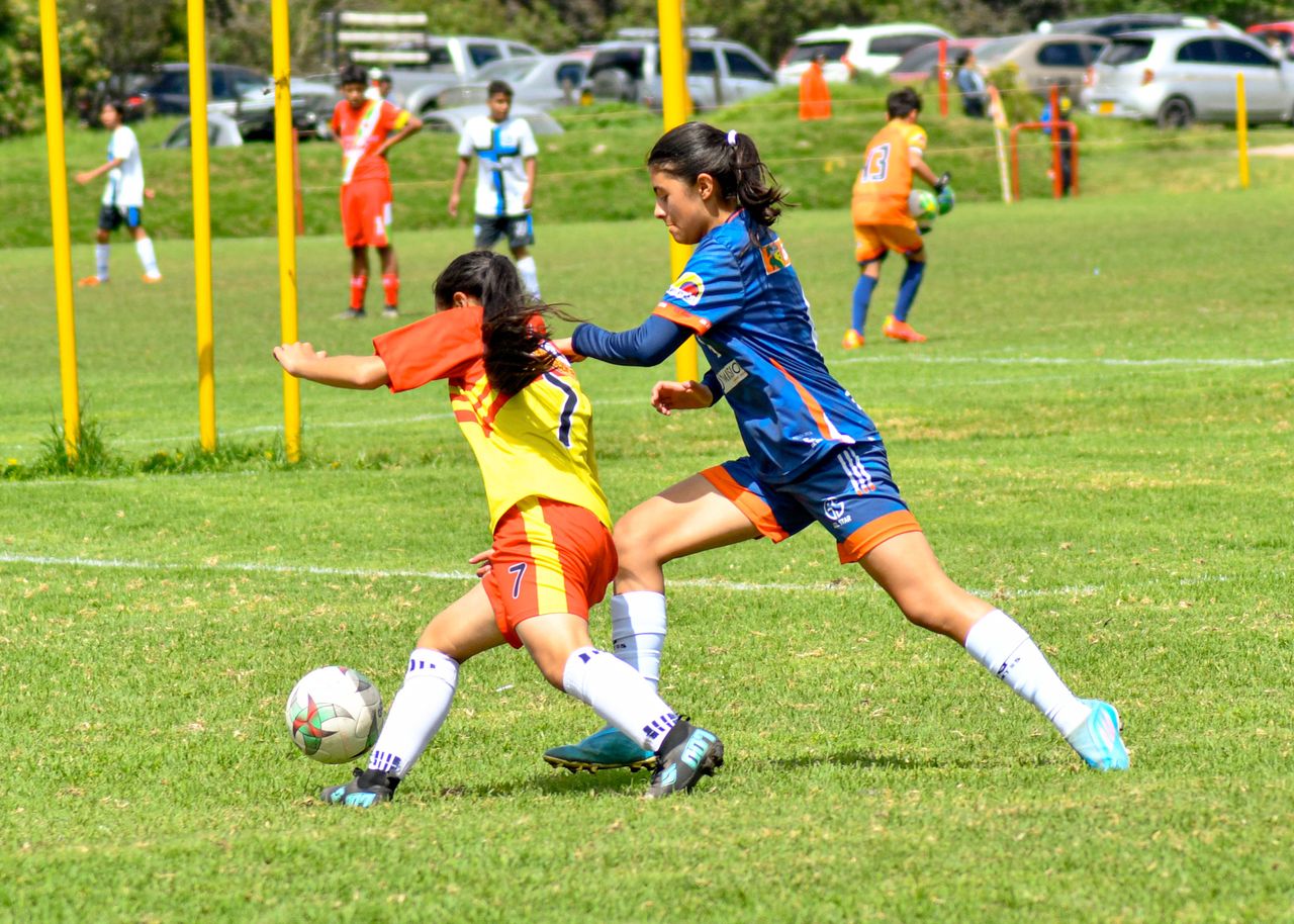 Two young girls playing football on a grass field, with other plays in motion in the background