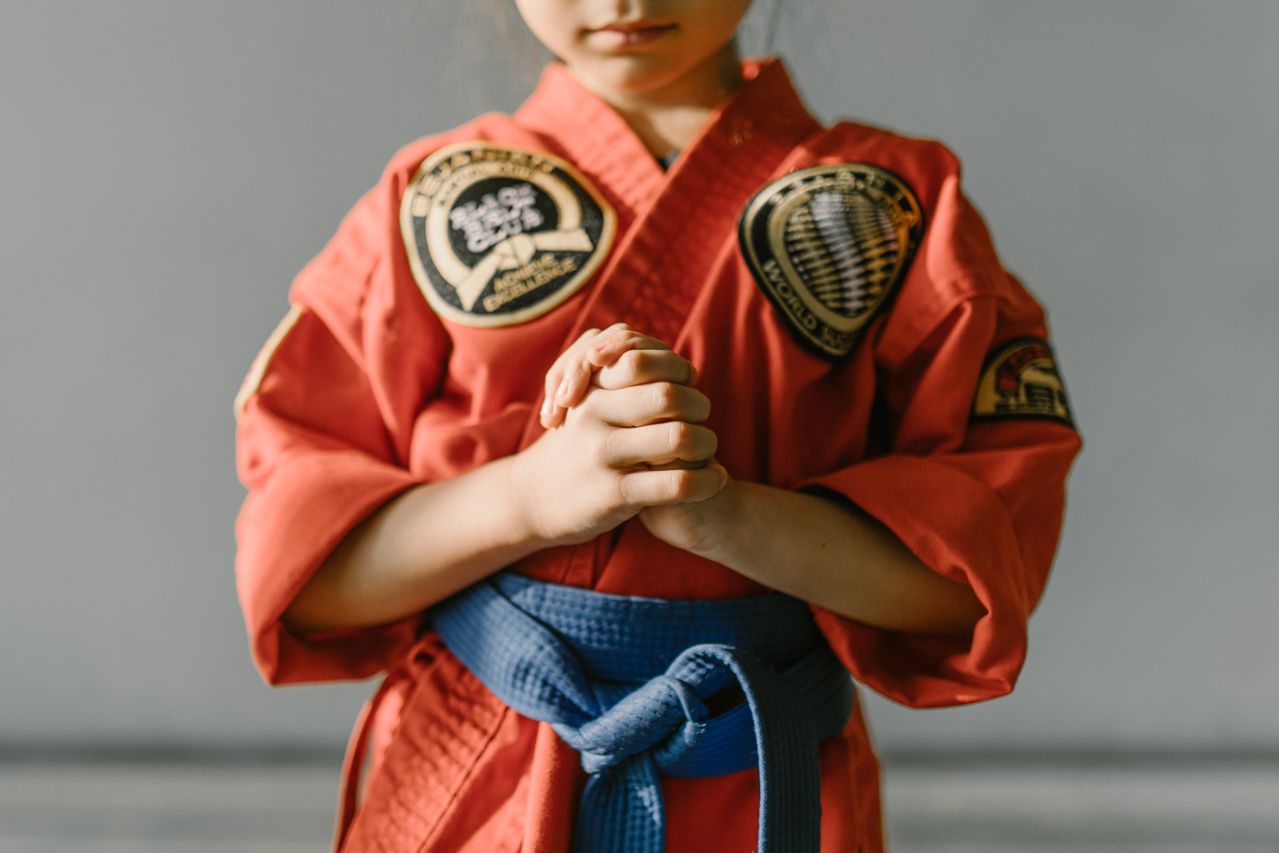 A young child in a red martial arts uniform with a blue belt, clasping hands in focus.