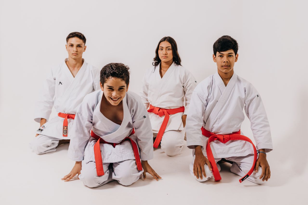 Four teenagers in martial arts uniforms with red belts against a white background.