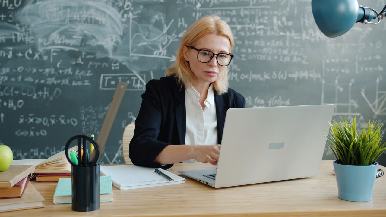 Teacher working on laptop with books and stationery around her on a table and a chalkboard full of equations in the background.