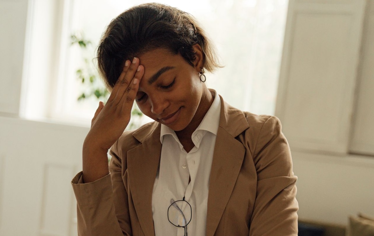 A professional woman stands in an office, appearing stressed or thoughtful, with a hand to her head.