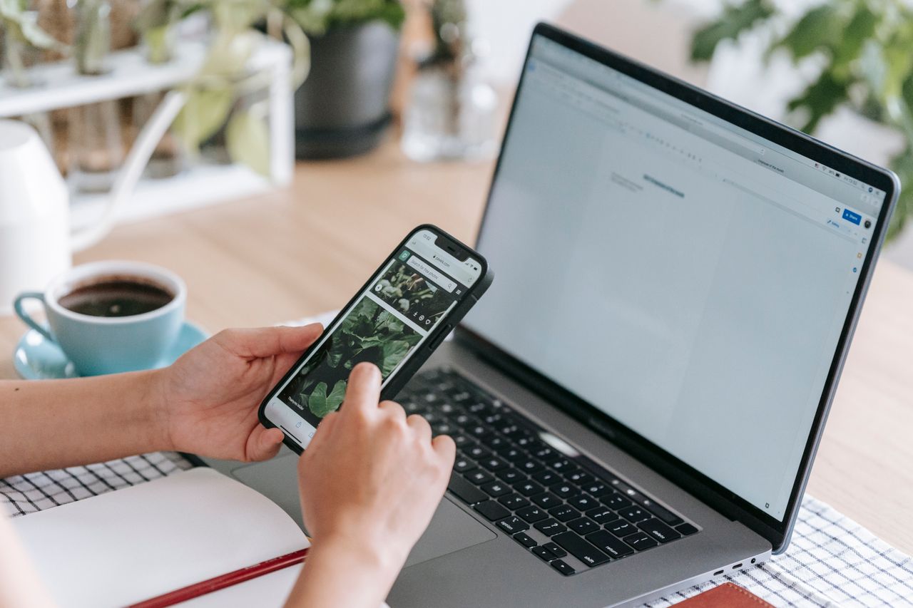 Person using a smartphone and laptop at a desk with coffee and plants.