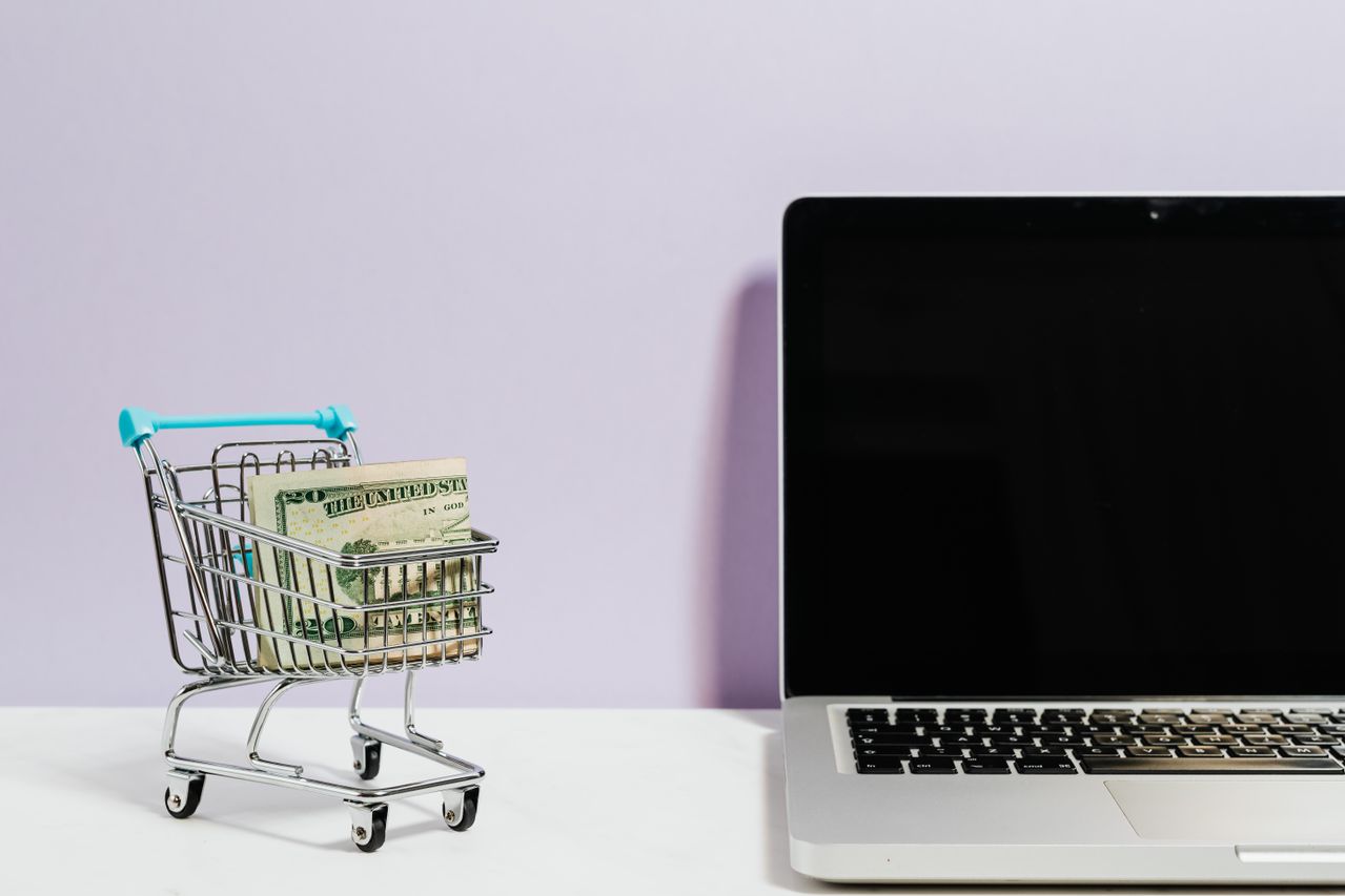 Shopping cart with money next to a laptop symbolizing online shopping.