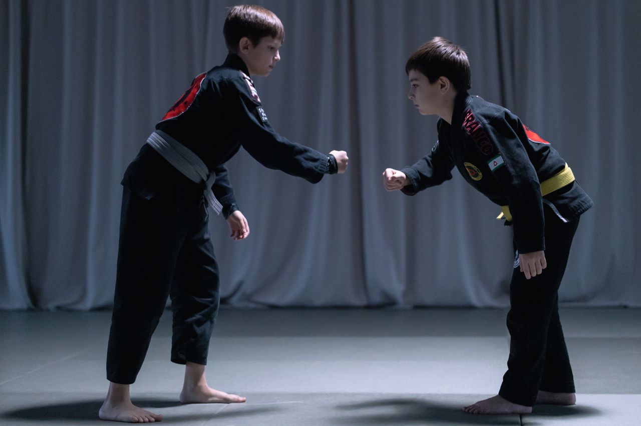 Two young boys in martial arts uniforms mirroring one another during a jiu jitsu practice session indoors.