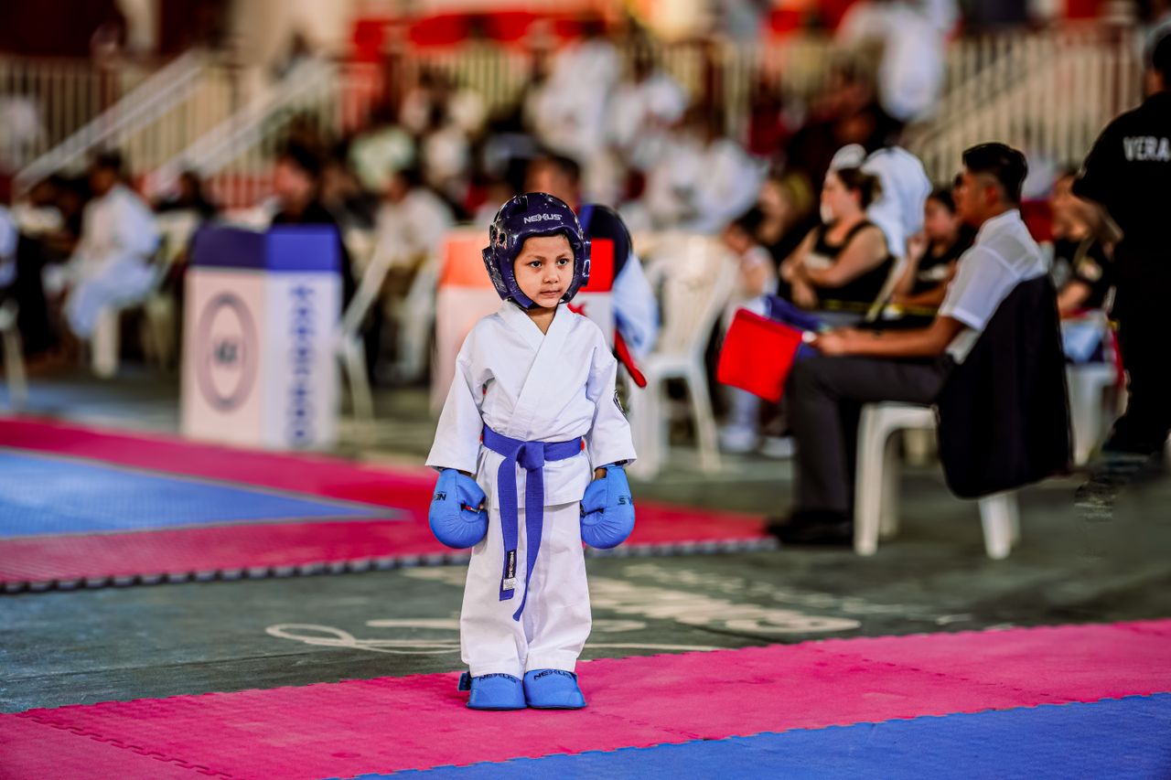A young karate competitor stands on a mat during a tournament, wearing blue protective gear.