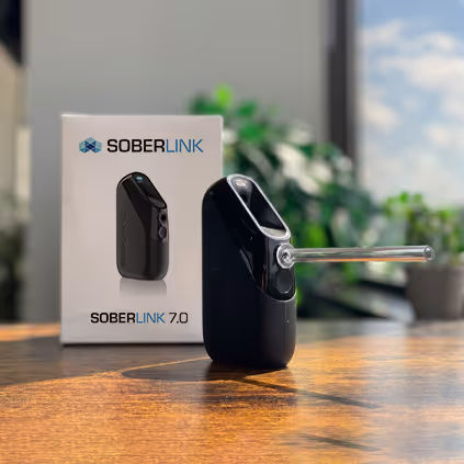 Soberlink 7.0 alcohol monitoring device with mouthpiece, placed on a wooden surface next to its white box packaging.