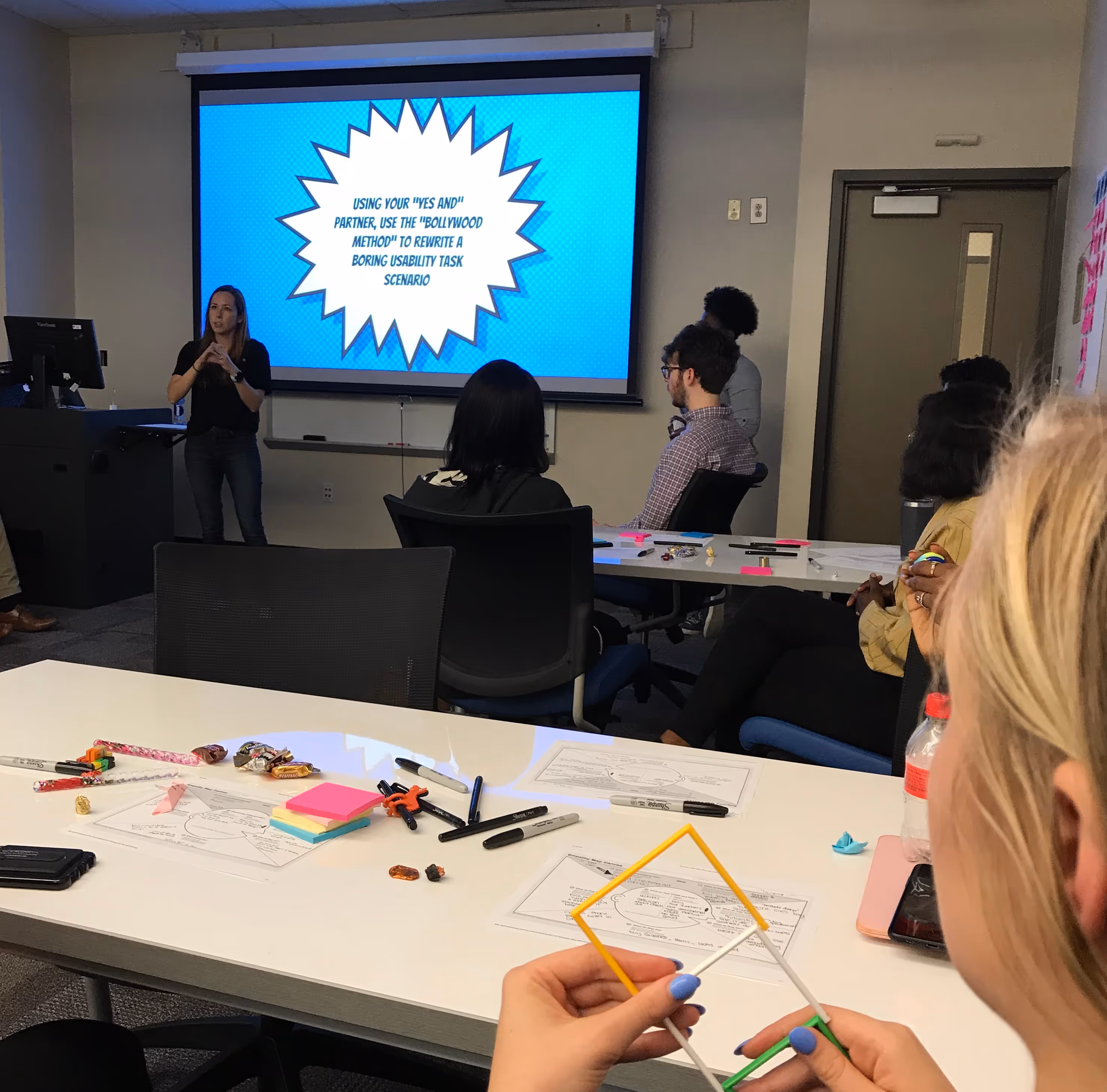 A image of a design workshop. A woman is standing in front and explaining design exercises to students.