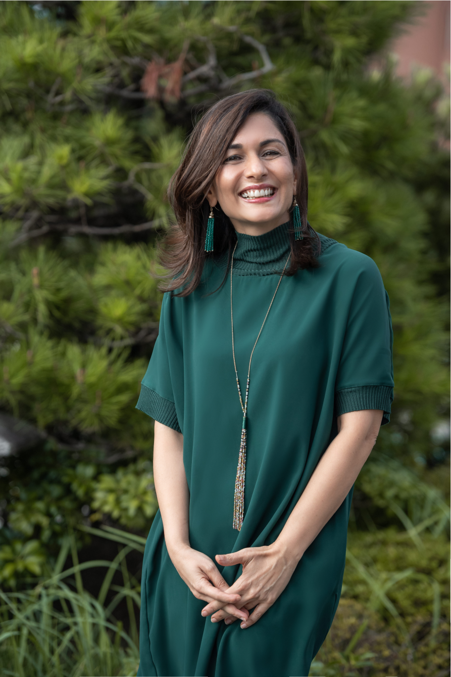 Smiling woman with dark hair wearing a green dress and long beaded necklace standing outdoors near greenery.