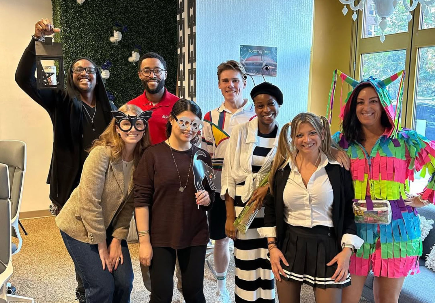 Group of eight people smiling indoors, wearing various fun costumes and accessories including a colorful piñata outfit, skull glasses, cat mask, and holding a lantern.