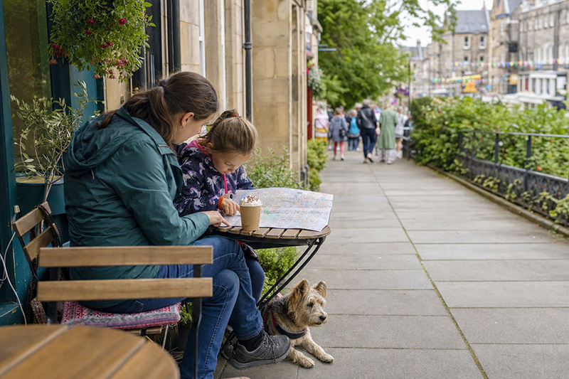 Parent and child checking a city map at an outdoor Edinburgh café table, with a dog resting beside them.