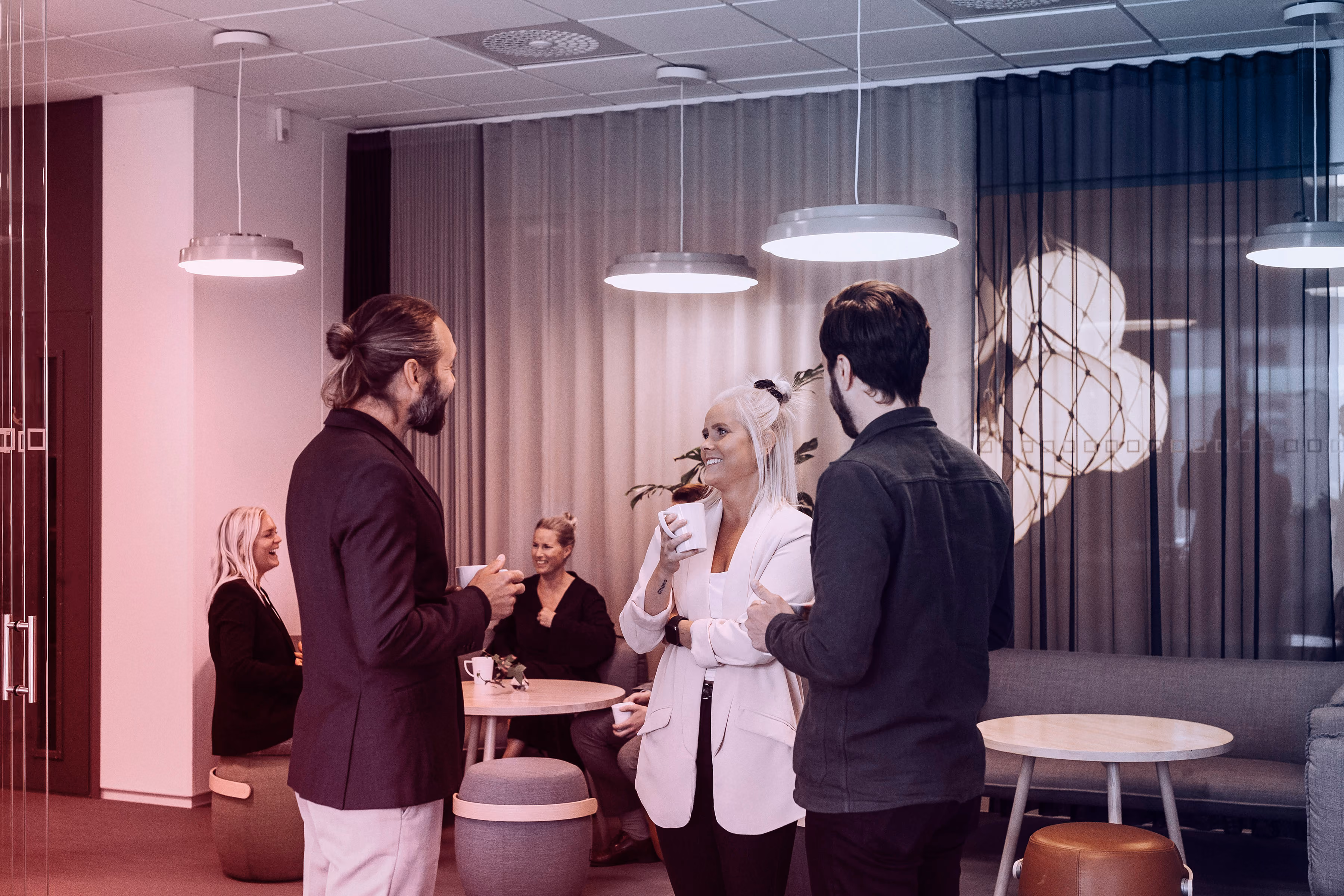 Drie mensen staan dicht bij elkaar en praten met koffiekopjes in een moderne kantoorruimte.