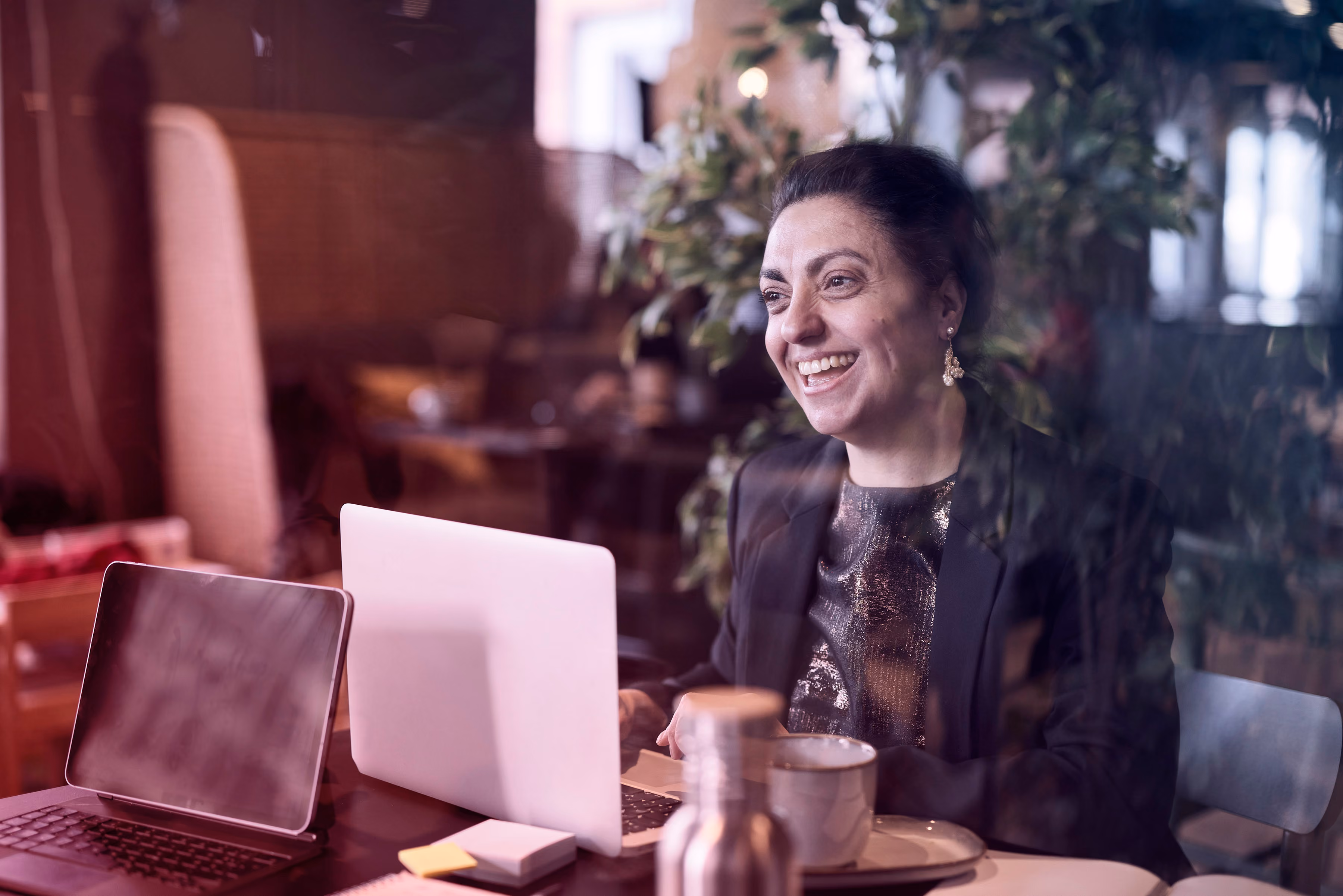 Lachende vrouw werkt op laptop in café met planten op de achtergrond.
