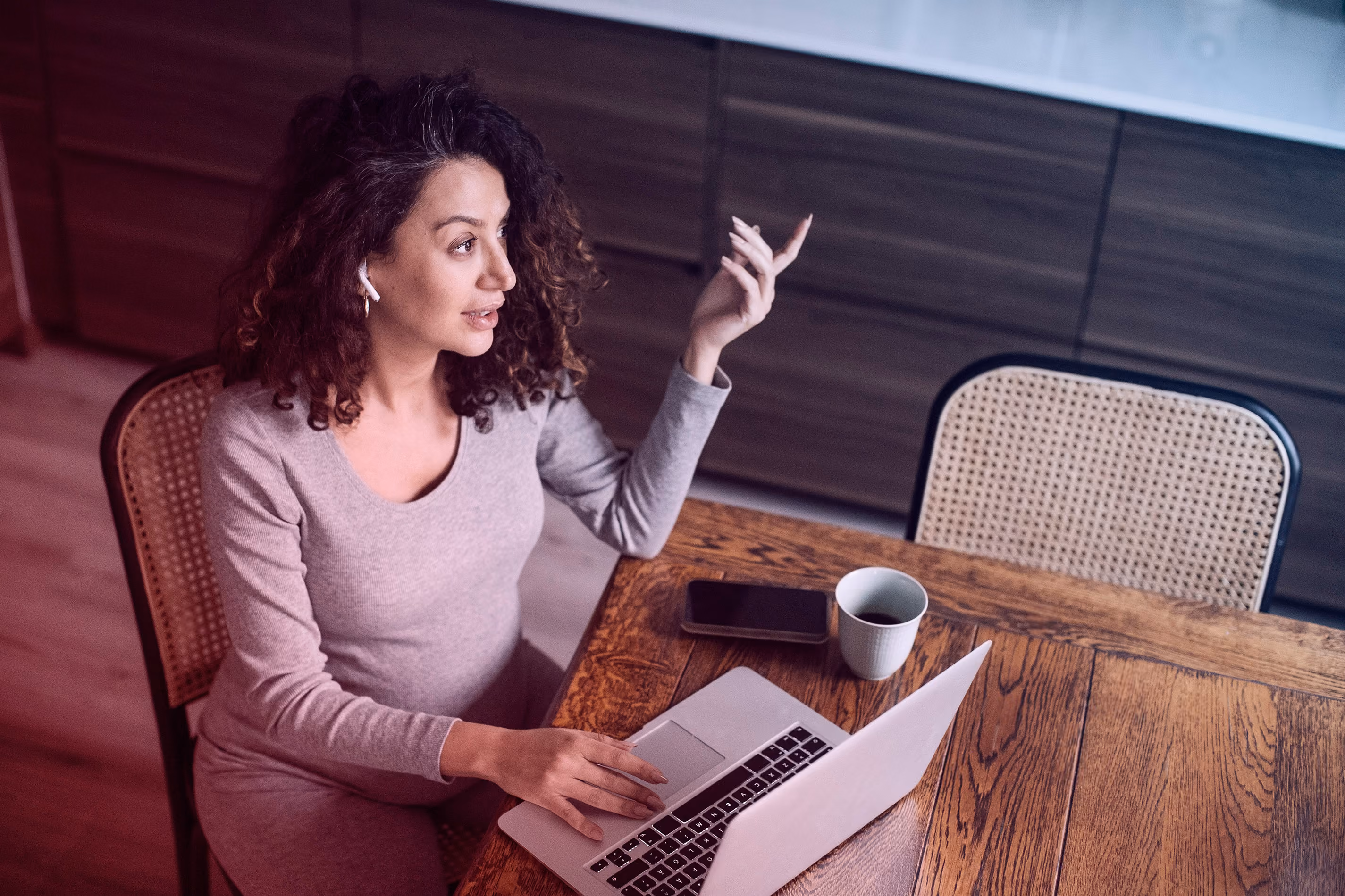 Vrouw met krullend haar werkt thuis op laptop met koffie en telefoon op hout tafel.