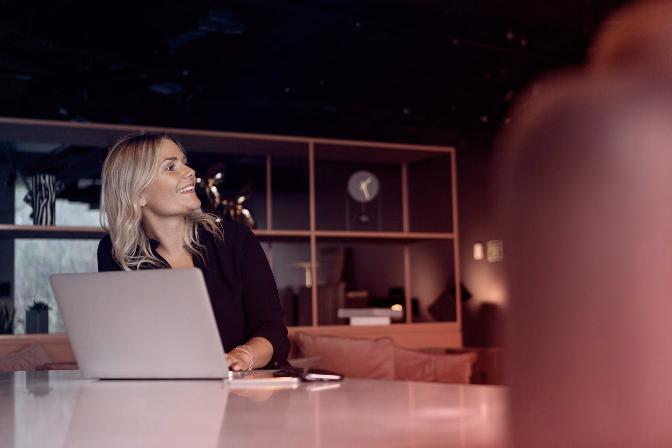 Vrouw met blond haar zittend achter laptop in een modern kantoor met boekenplank en klok.