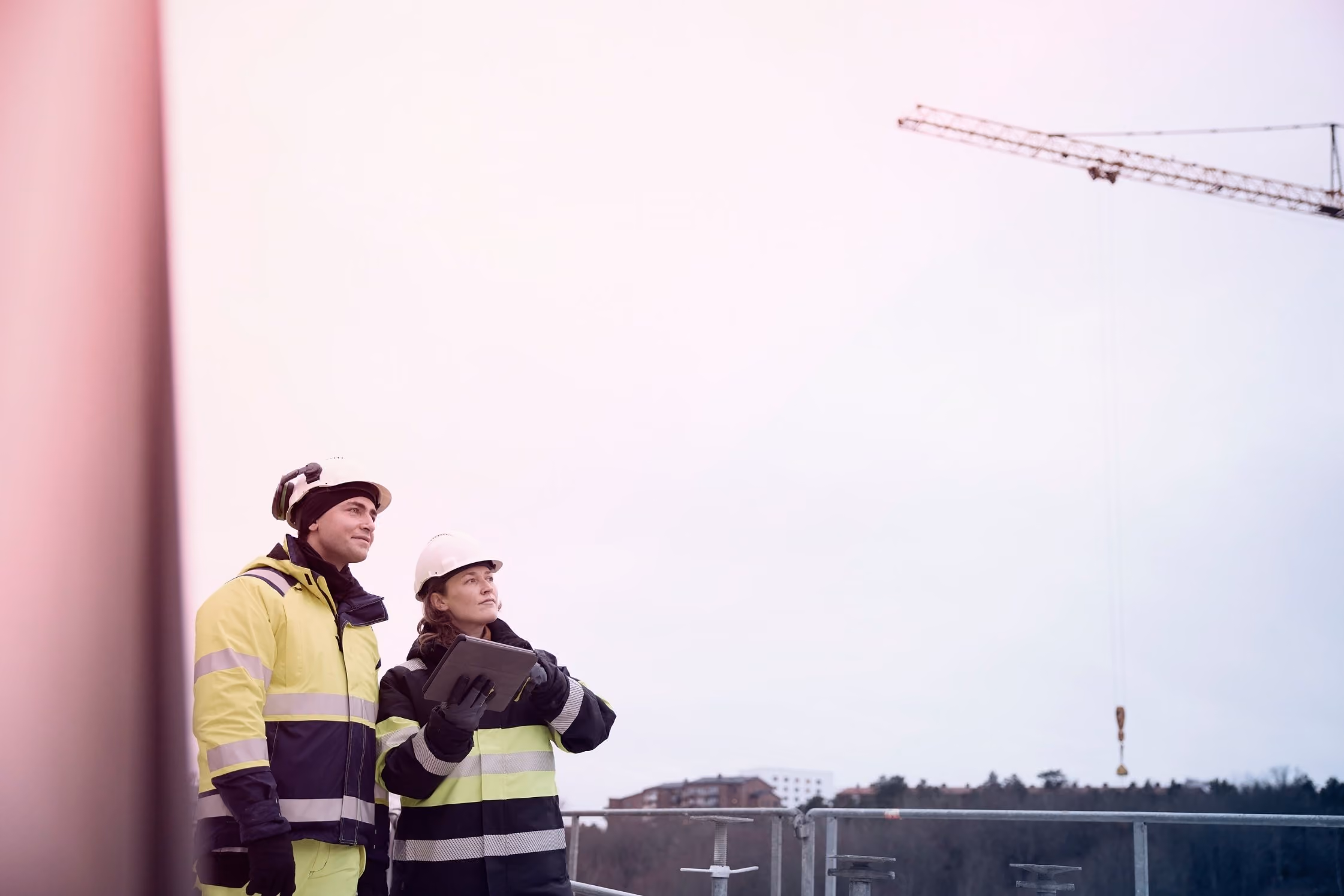 Twee bouwvakkers in veiligheidshesjes en helmen bekijken een bouwkraan buiten op een bouwplaats.