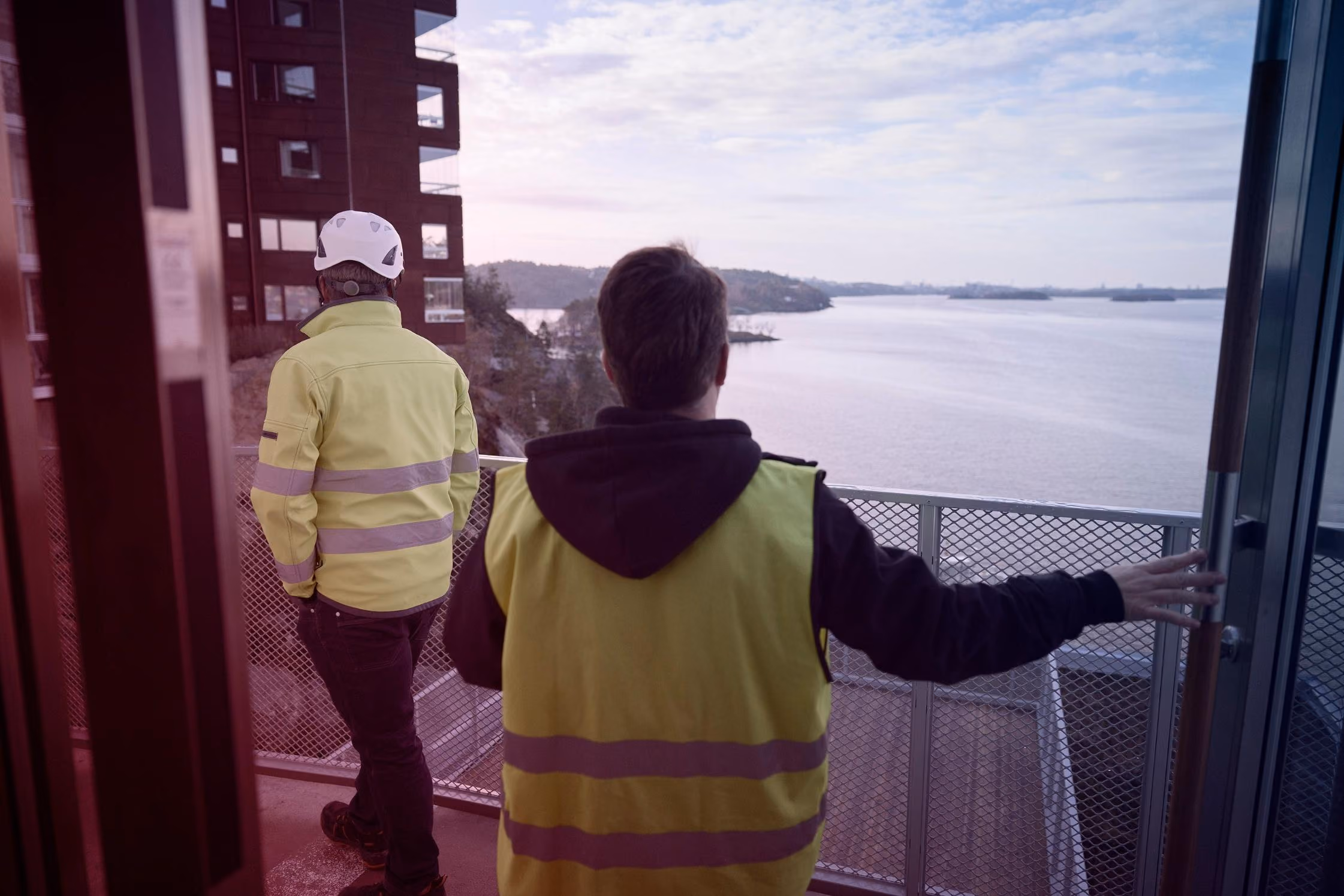 Twee bouwvakkers in geel veiligheidshesje kijken uit over water vanaf een balkon.