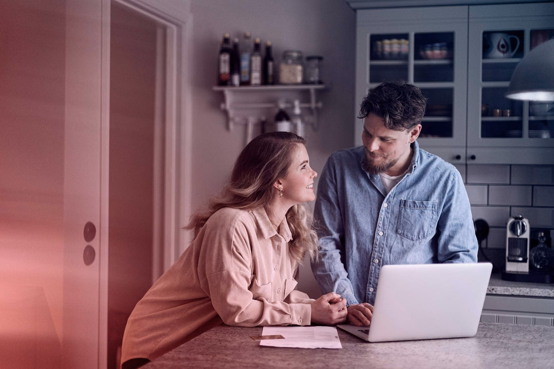 Man en vrouw praten lachend bij laptop in de keuken, met documenten op het aanrecht.