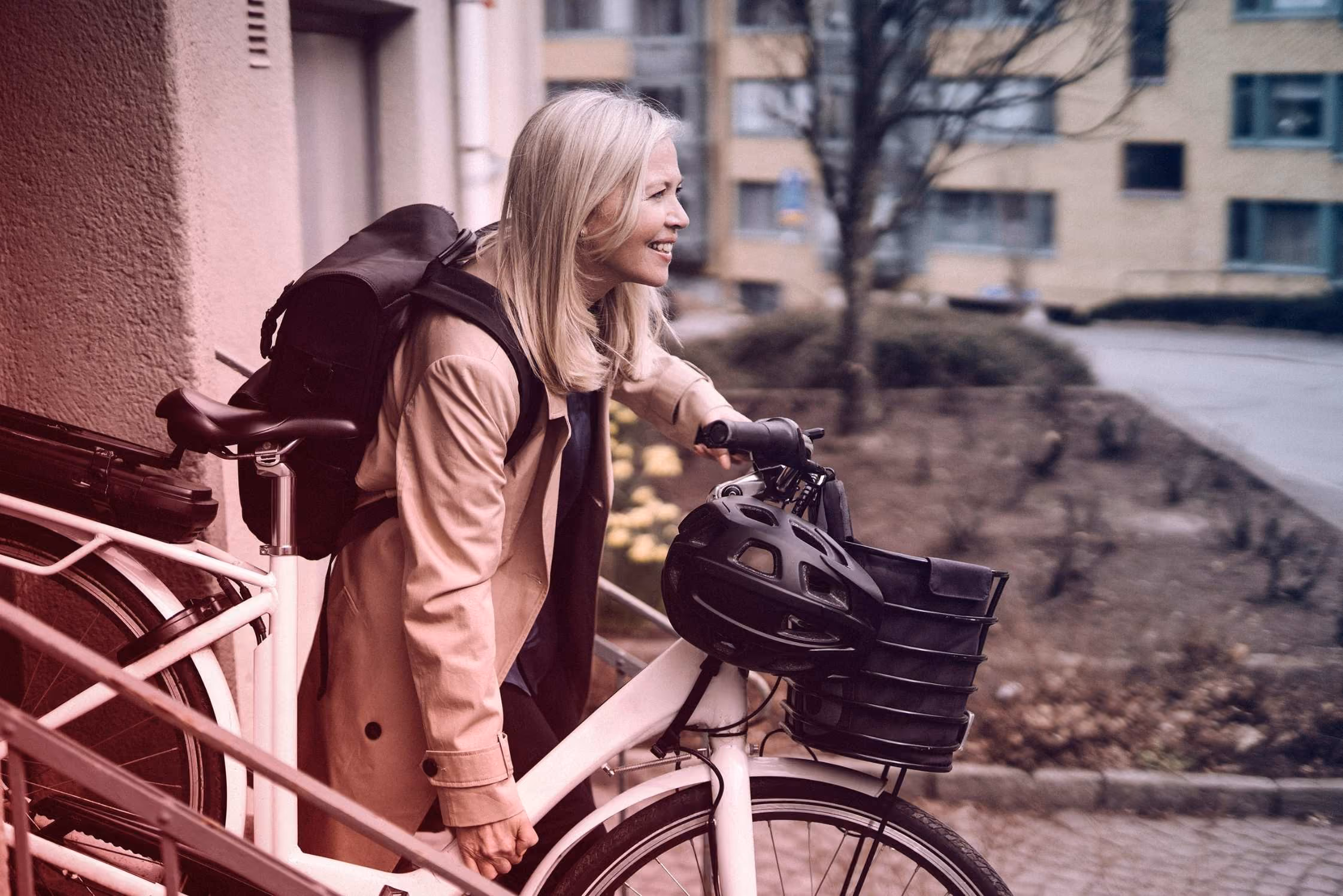Vrouw met licht haar, beige jas en rugzak, duwt een witte fiets met zwarte helm in een stedelijke omgeving.