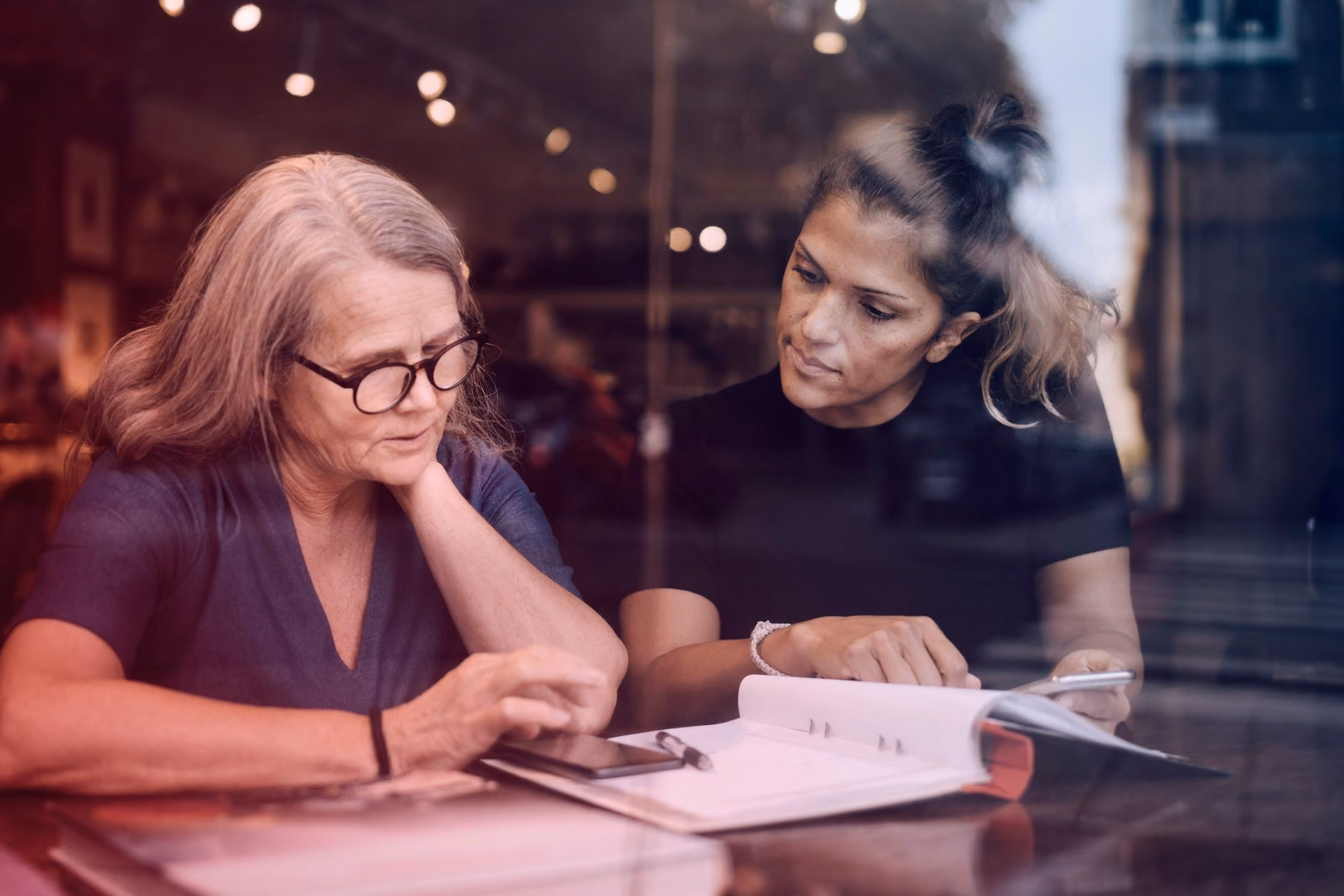 Twee vrouwen bespreken documenten en gebruiken een smartphone aan een tafel in een café.