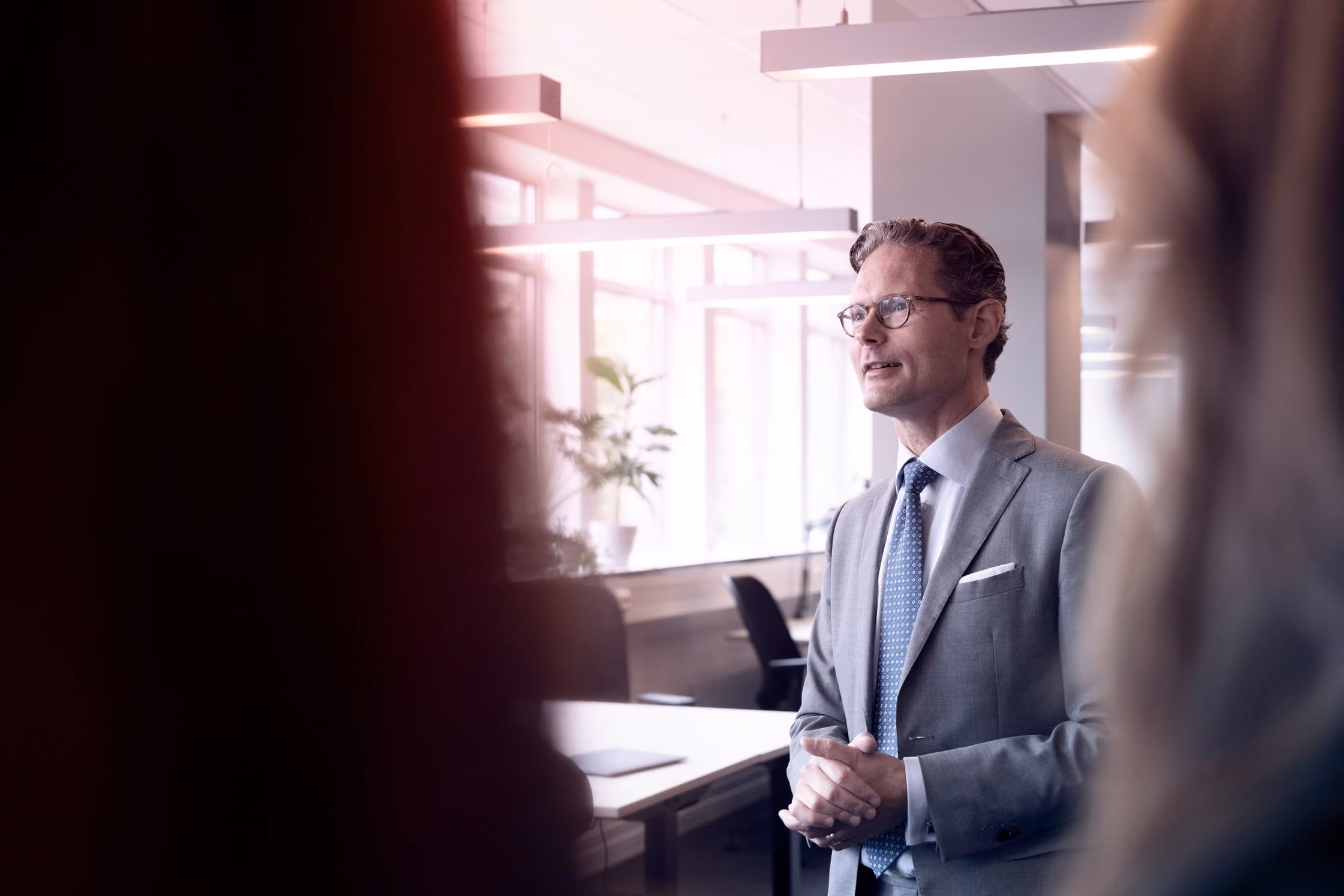 Zakelijke man in pak en bril spreekt in een modern kantoor met veel licht en bureaus.