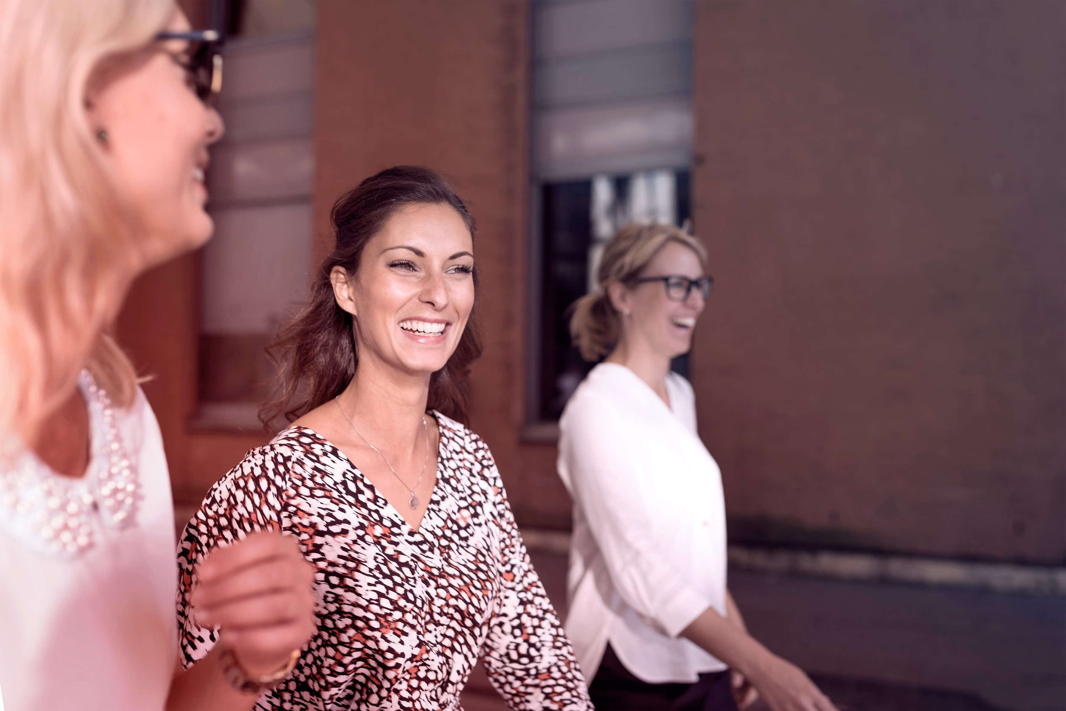 Drie vrouwen die buiten lopen en lachen, gekleed in casual kleding, tegen een achtergrond van een roodbruine bakstenen muur met een raam.