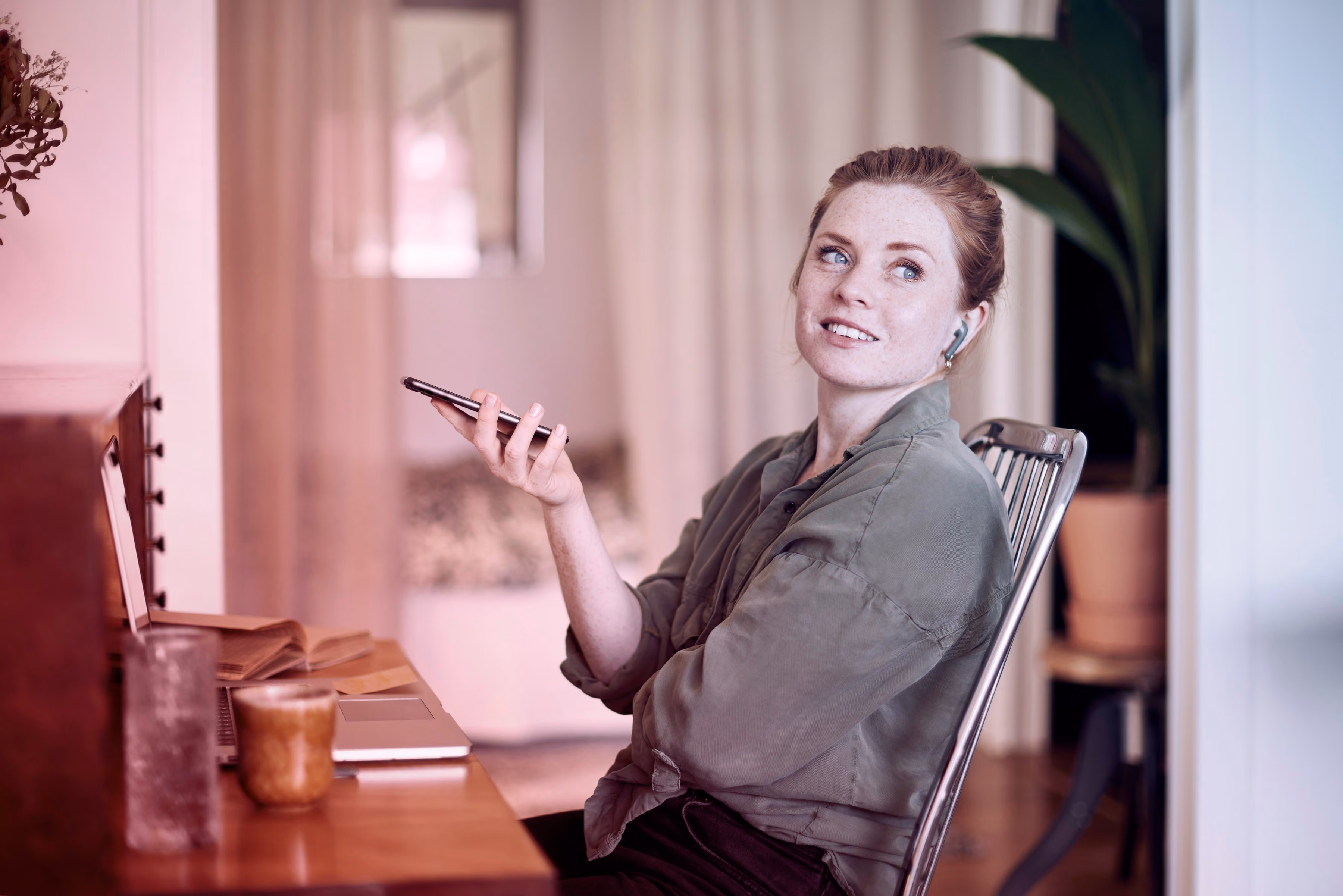 Vrouw met oordopjes draagt een groen jasje en zit op een stoel aan een houten bureau met een laptop, mok en glazen op de voorgrond, terwijl ze naar rechts kijkt en een smartphone in haar hand houdt.