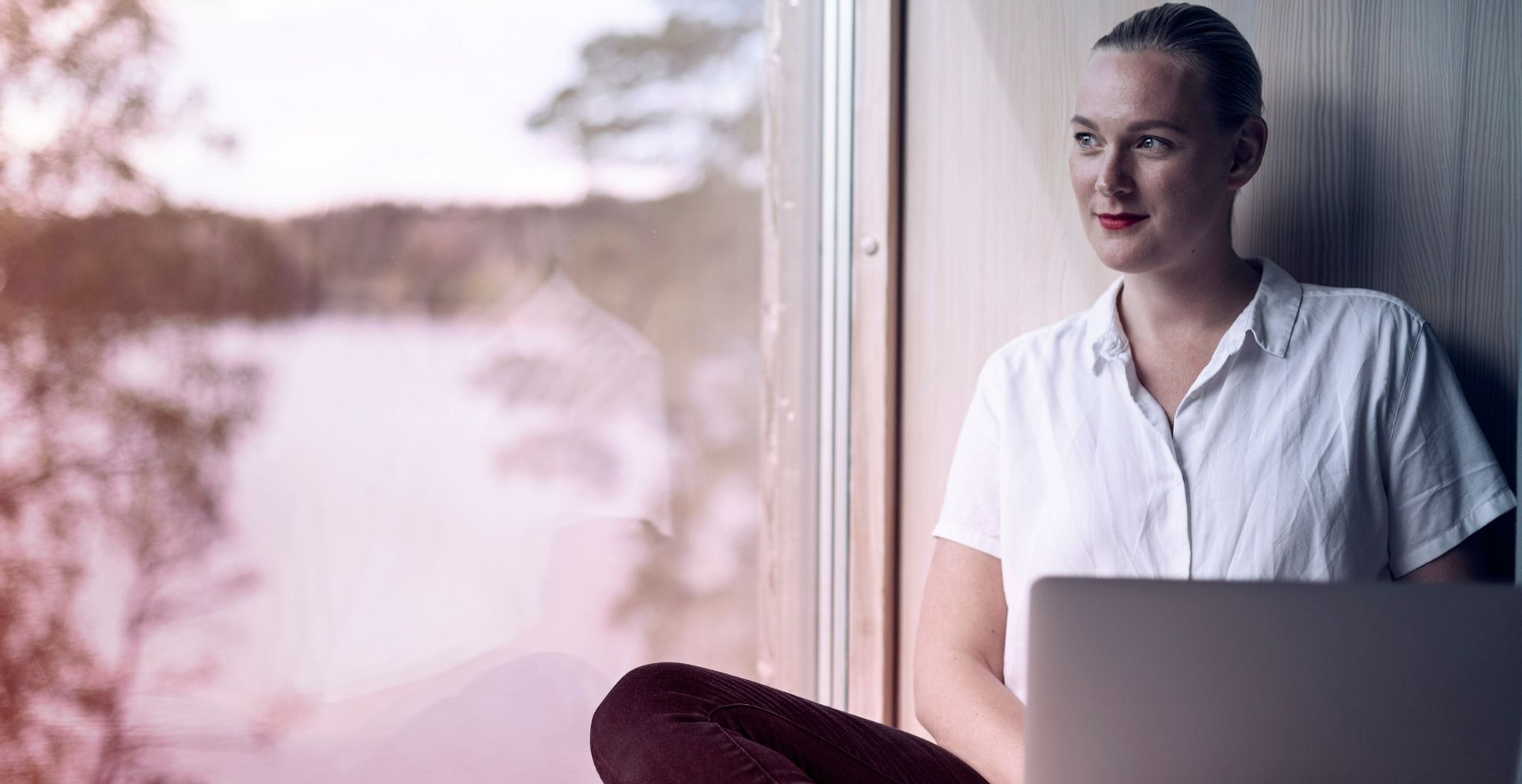Vrouw zit met laptop op schoot voor raam en kijkt naar buiten