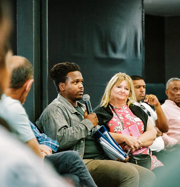 A seated man speaking to a seated audience, who are engaged and listening attentively in a meeting setting.