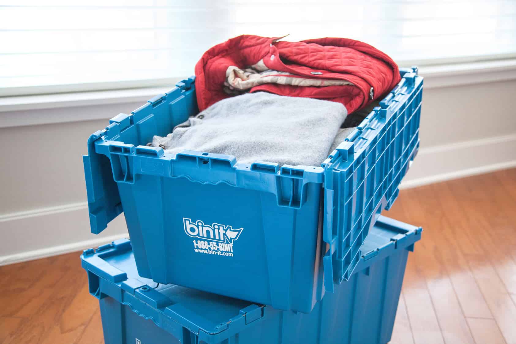 Folded clothes packed in a blue Bin It bin, stacked on a wood floor