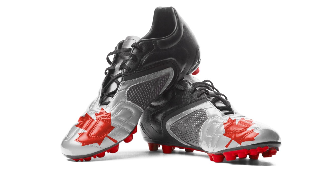 A pair of black and silver soccer cleats with red studs. Each shoe features a large red maple leaf graphic on the front, symbolizing Canada. The cleats are crossed and positioned against a white background, with one shoe resting on top of the other.