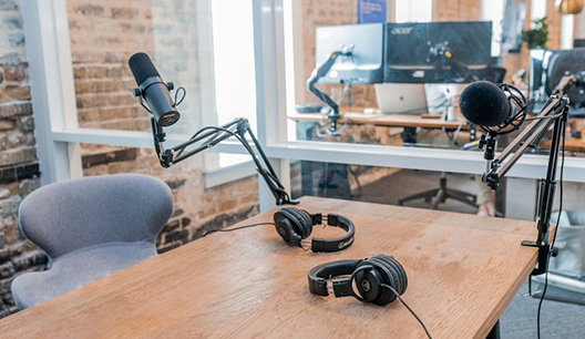 Podcast recording setup with two microphones on adjustable arms and two headphones on a wooden table in a modern office.
