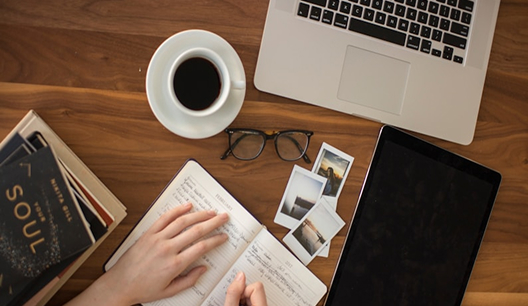 Person writing in a notebook on a wooden desk with a cup of coffee, eyeglasses, a laptop, a tablet, three photos, and books nearby.