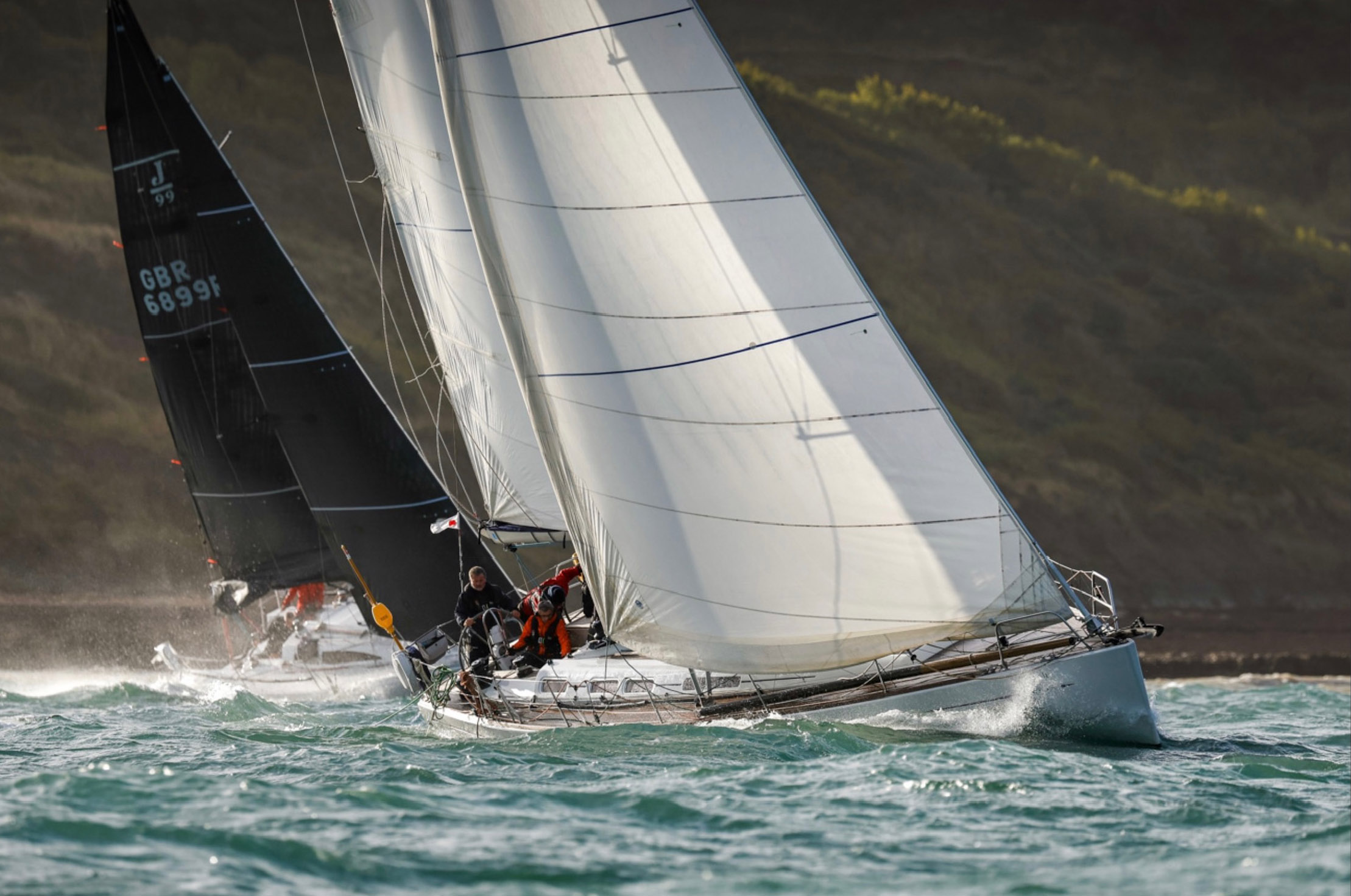 Two sailboats racing on choppy water near a green, rocky coastline under a partly cloudy sky.