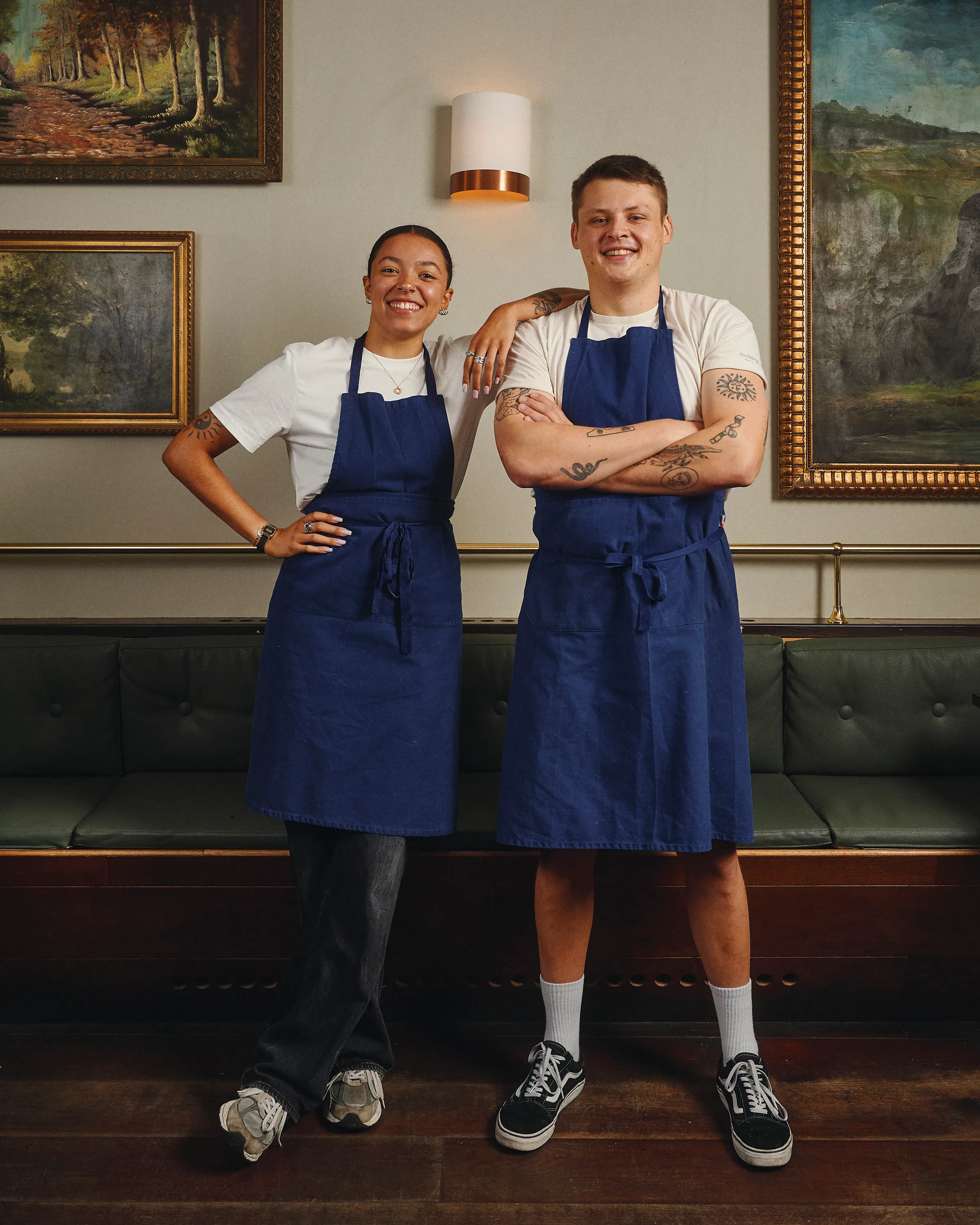Two smiling people wearing blue aprons and white shirts standing indoors with framed paintings on the wall behind them.