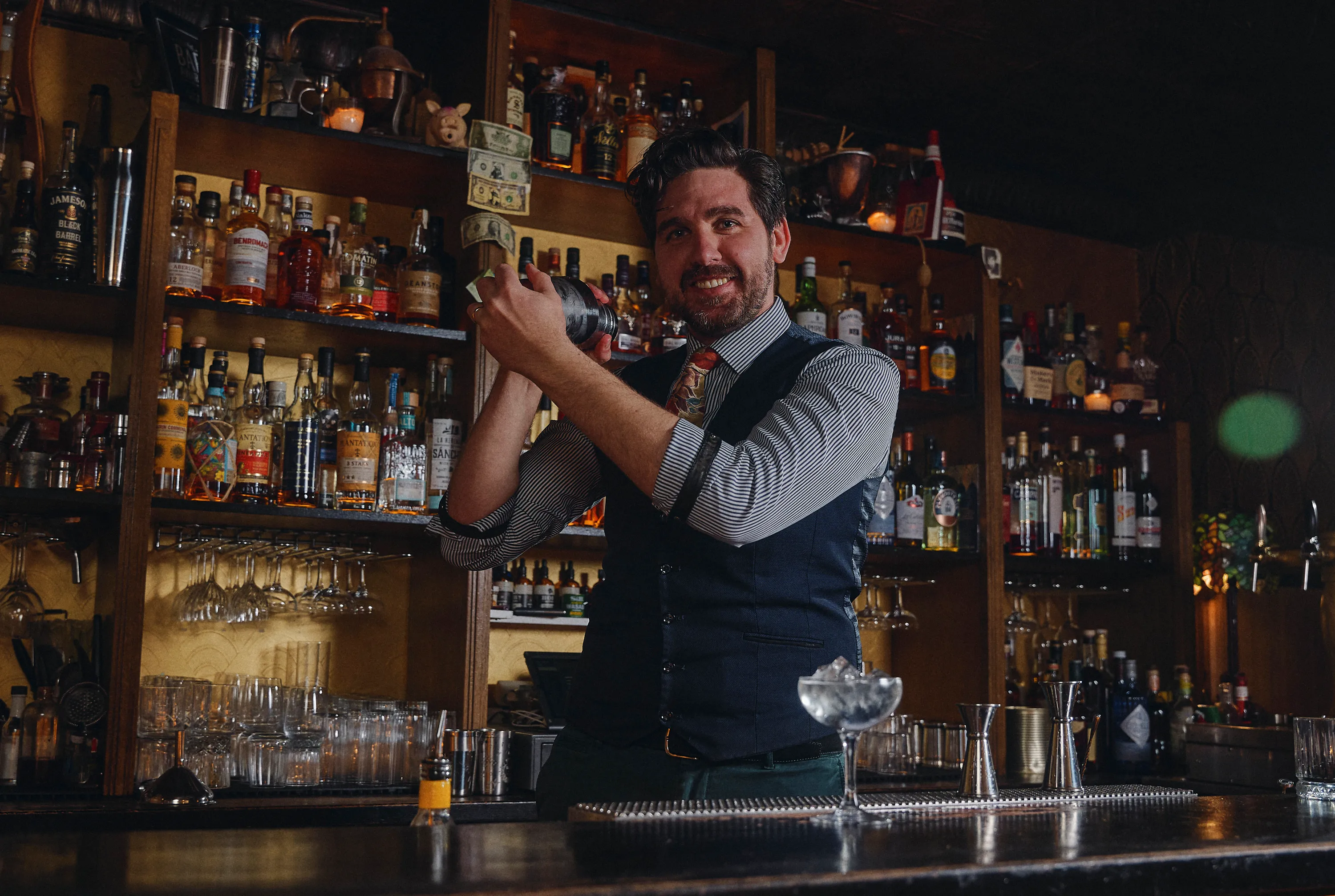 Smiling bartender shaking a cocktail shaker behind a bar with shelves of liquor bottles and glassware.