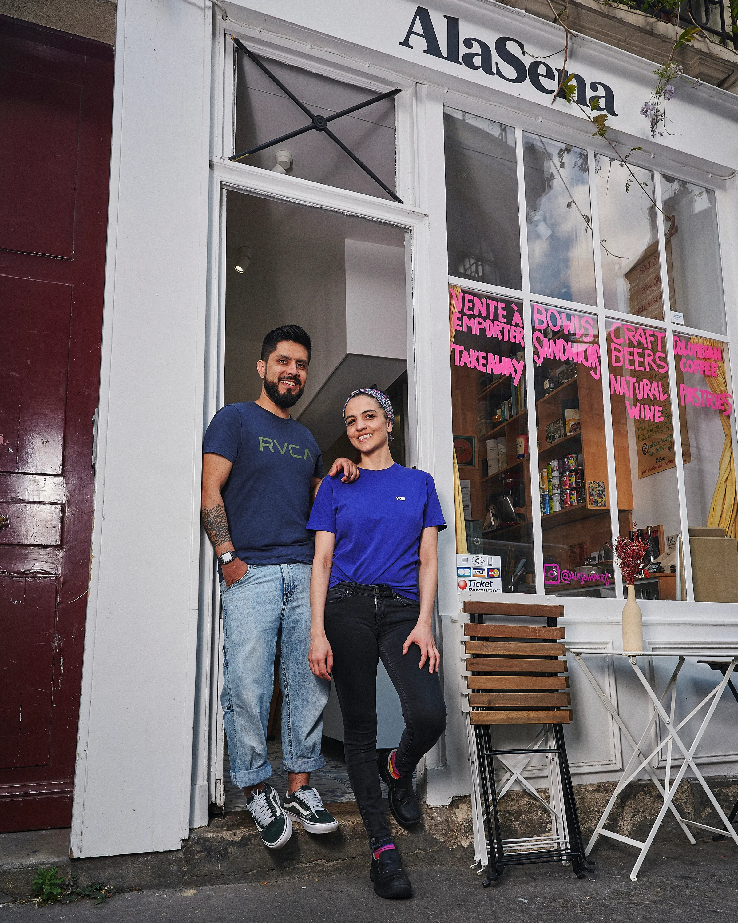 Two smiling people standing in the doorway of AlaSena café with menu items written in pink on the window.