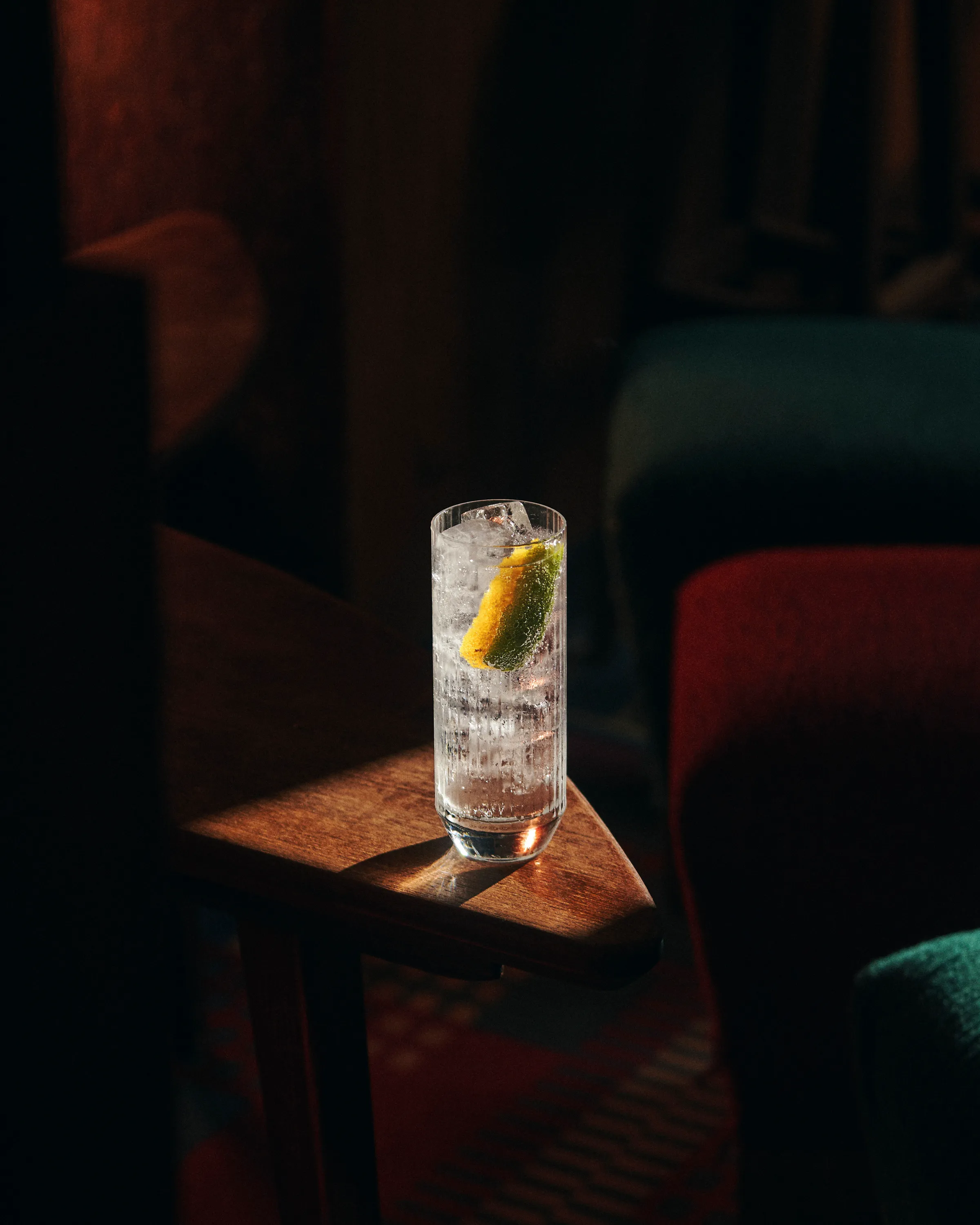 Tall glass with ice and a lemon wedge on a wooden armrest in dim lighting.
